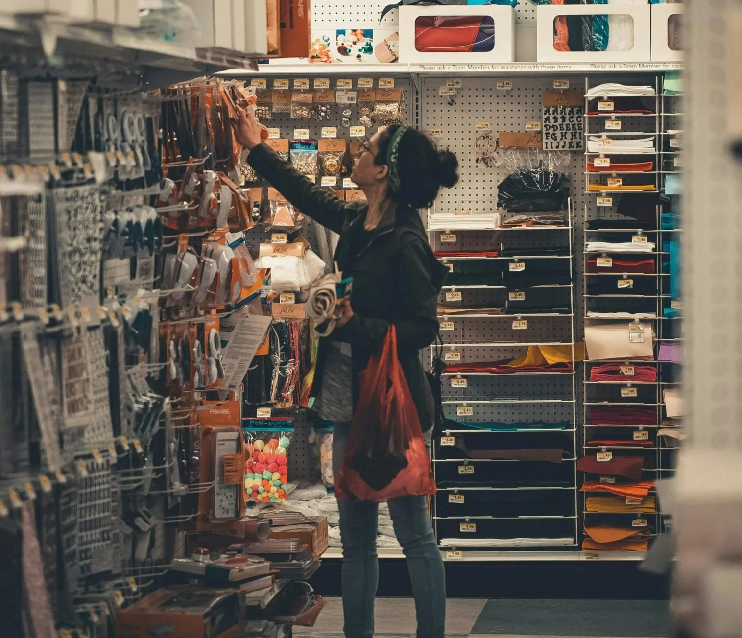 A woman shopping in a craft store, reaching for an item on a shelf filled with craft supplies.