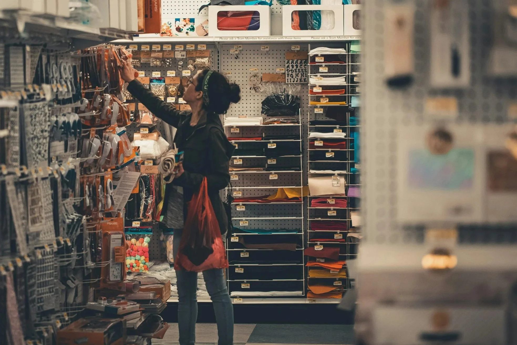 A woman shopping in a store aisle with craft supplies, reaching for an item on the shelf, surrounded by organized shelves of paper, folders, and stationery.