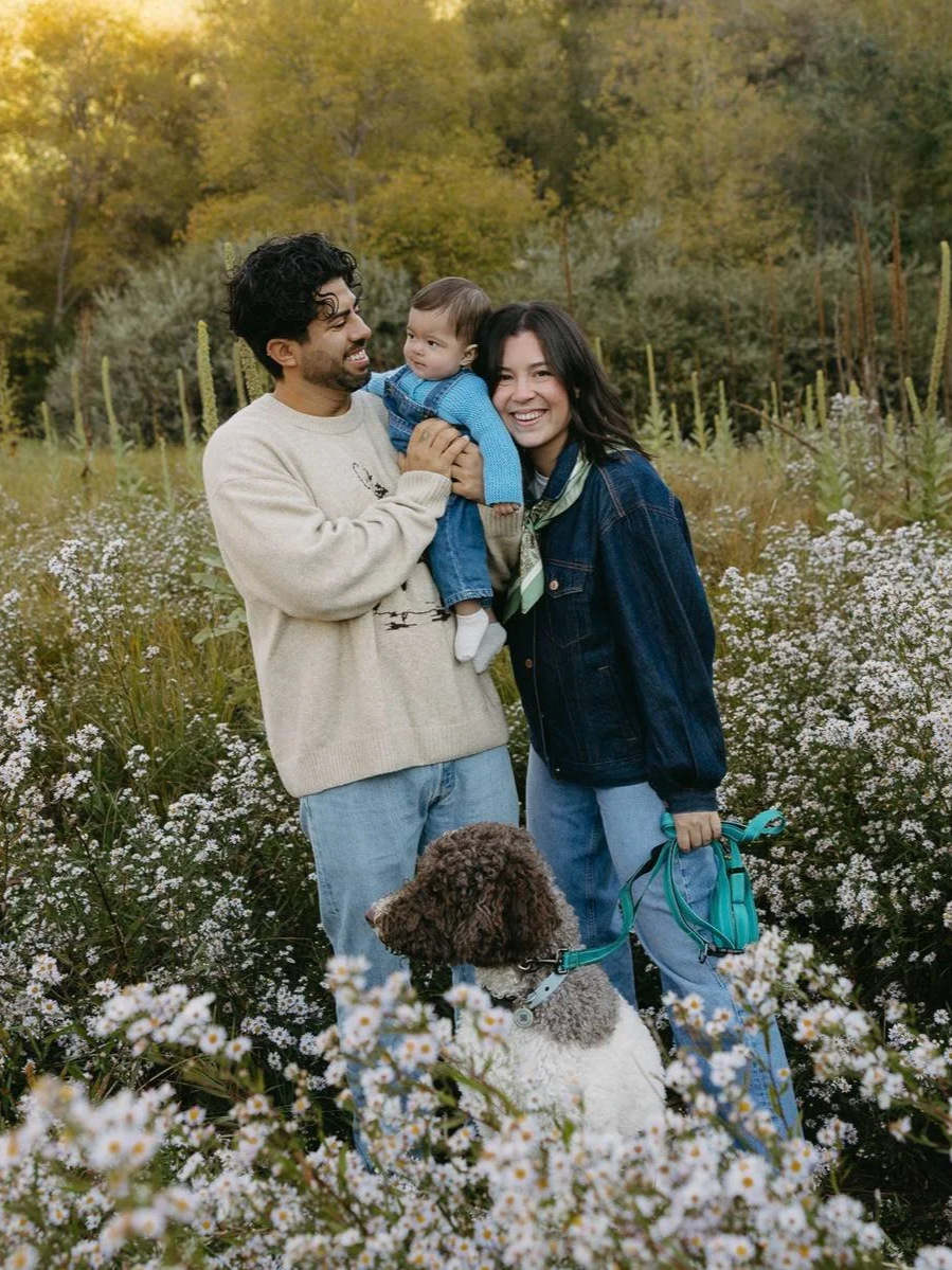 A happy family of three with a dog in a field of flowers and trees, during autumn, smiling and enjoying each other's company.