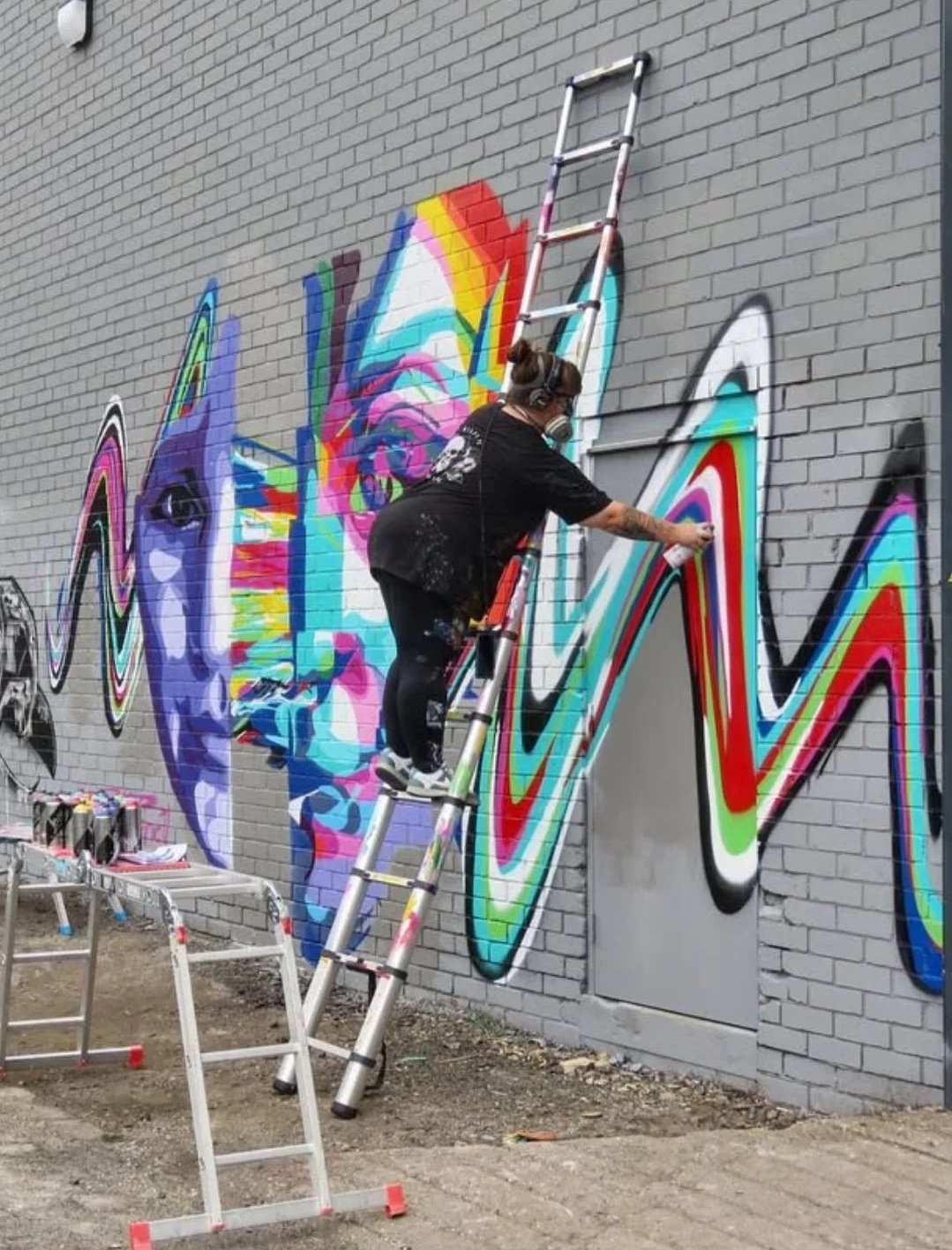 An artist is spray painting a colorful graffiti mural on a brick wall. The artist is on a ladder, wearing a black shirt and a respirator mask, working on vibrant, swirling abstract patterns.