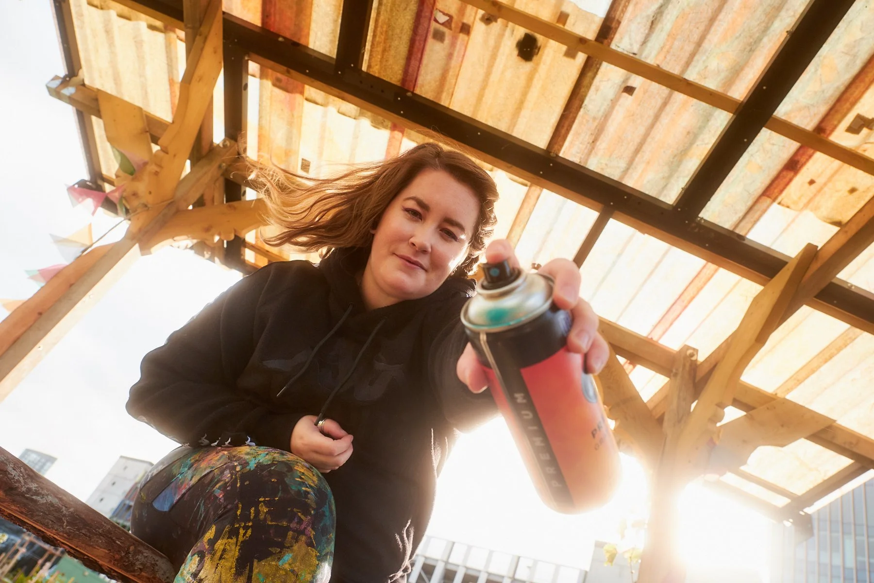 A young woman with shoulder-length hair, wearing a black hoodie and colorful artist pants, is holding and spraying paint from a spray can towards the camera. She is standing under a wooden pergola with a partly cloudy sky and tall buildings in the background.
