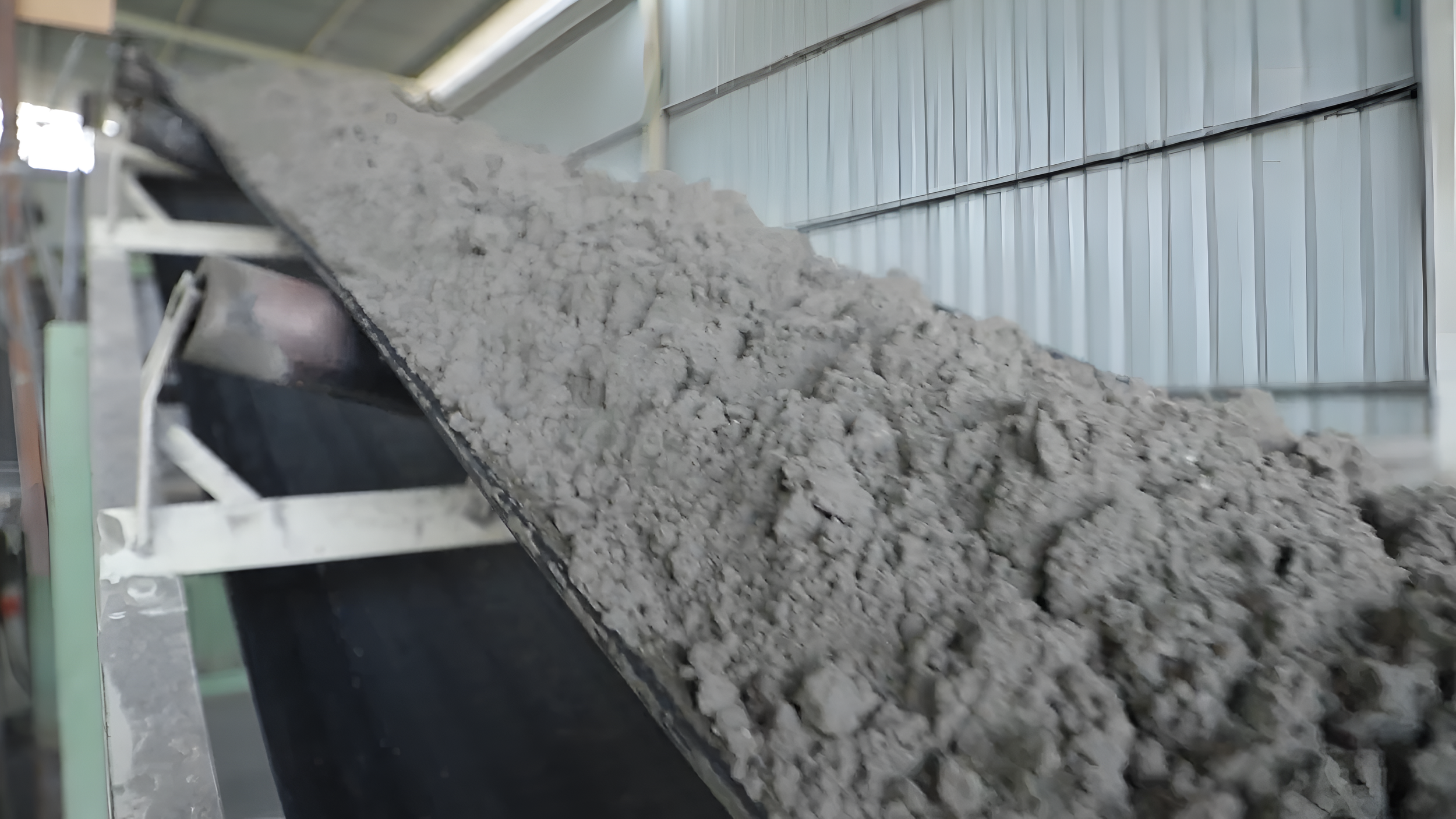 Conveyor belt transporting a large pile of gray material, possibly cement or similar powdery substance, inside an industrial building with metal walls and a high ceiling.