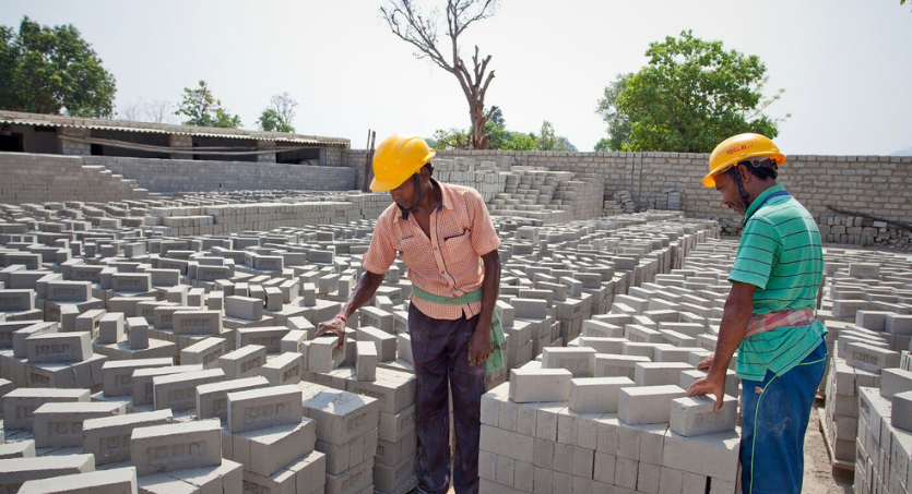 Two construction workers wearing yellow safety helmets inspecting a large area of stacked bricks at a construction site outdoors.