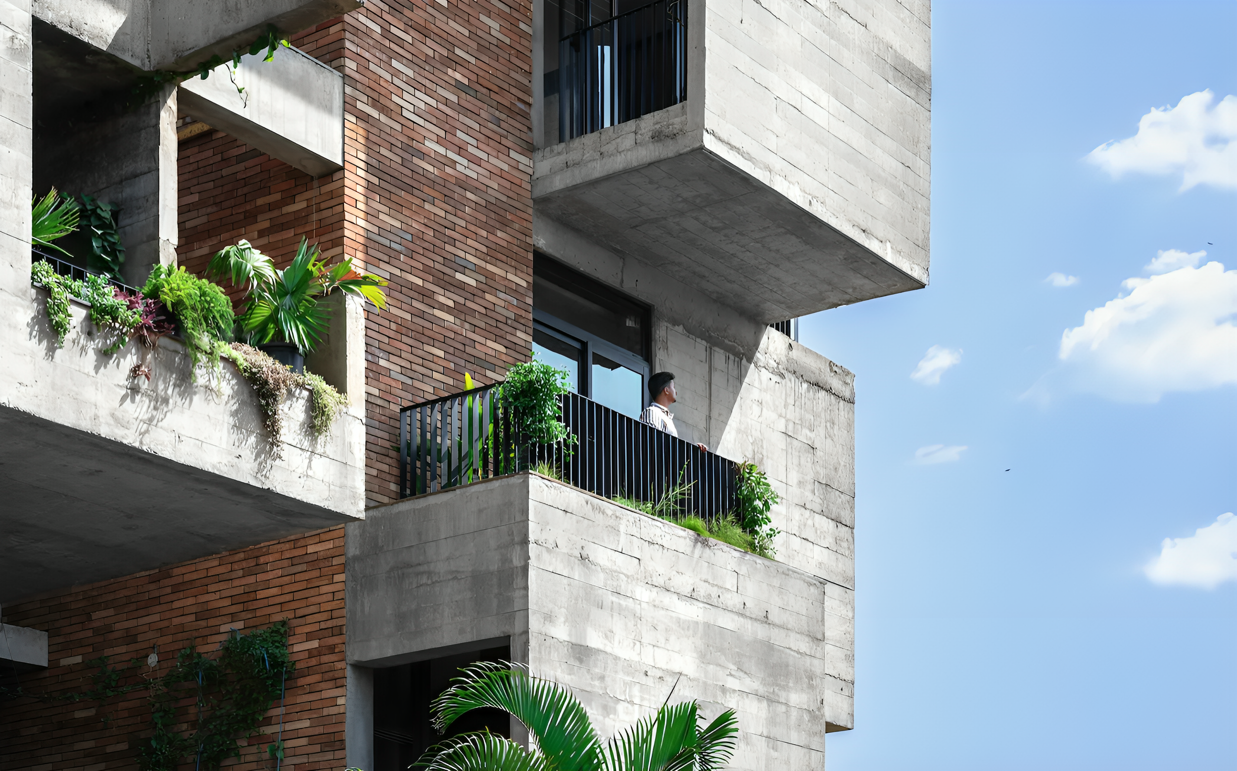 A modern apartment building with concrete, brick, and metal accents. A person is standing on a balcony small garden with plants, under a clear blue sky.