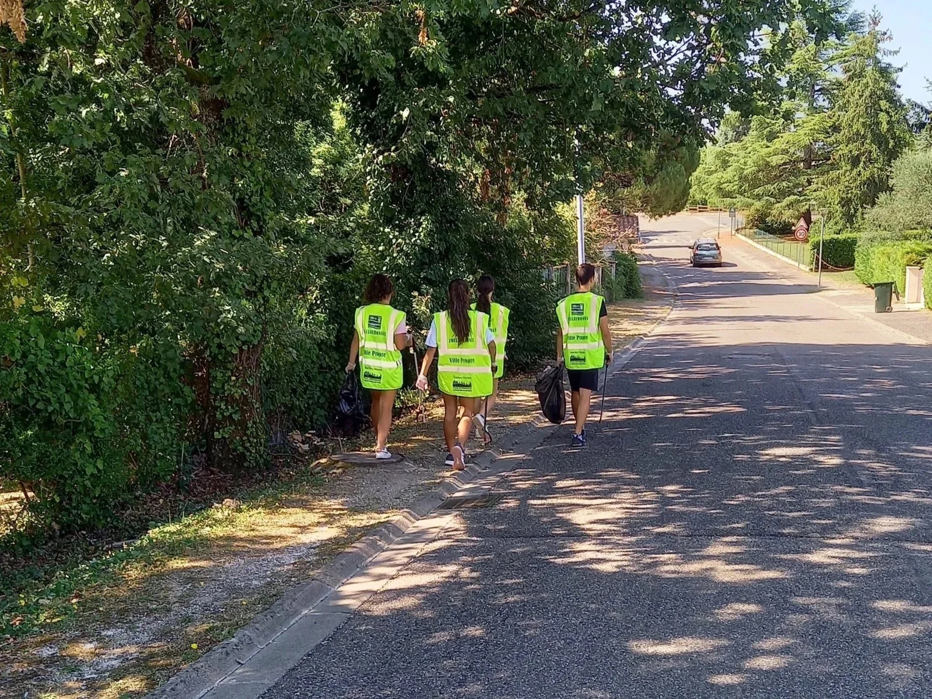 Groupe de quatre personnes portant des vestes jaunes fluorescentes avec des textes, marchant sur le trottoir ombragé d'un quartier résidentiel avec des arbres et des voitures au loin.