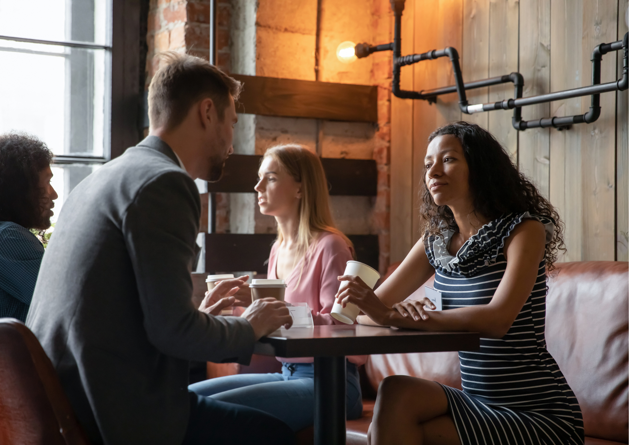 Quatre jeunes adultes discutent et prennent un café dans un café, assis autour d'une table en bois contre un mur en bois avec des éléments métalliques et une grande fenêtre.