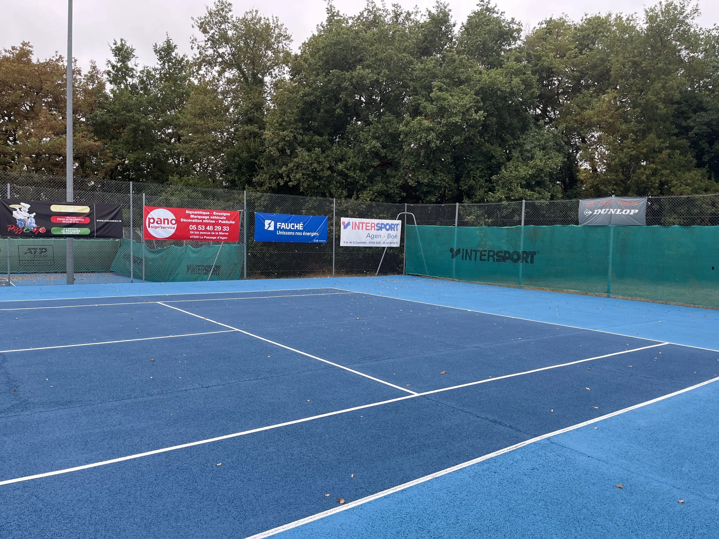 Un court de tennis en pleine nature avec un sol bleu et des filets blancs, entouré de clôtures et de panneaux publicitaires.