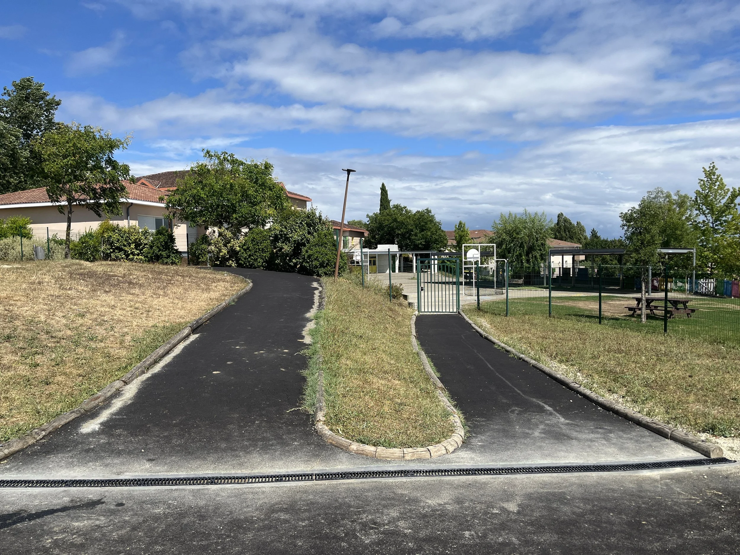 Un chemin pavé mène vers une aire de jeux pour enfants avec une clôture verte, dans un parc, avec des maisons en arrière-plan, des arbres et un ciel partiellement nuageux.