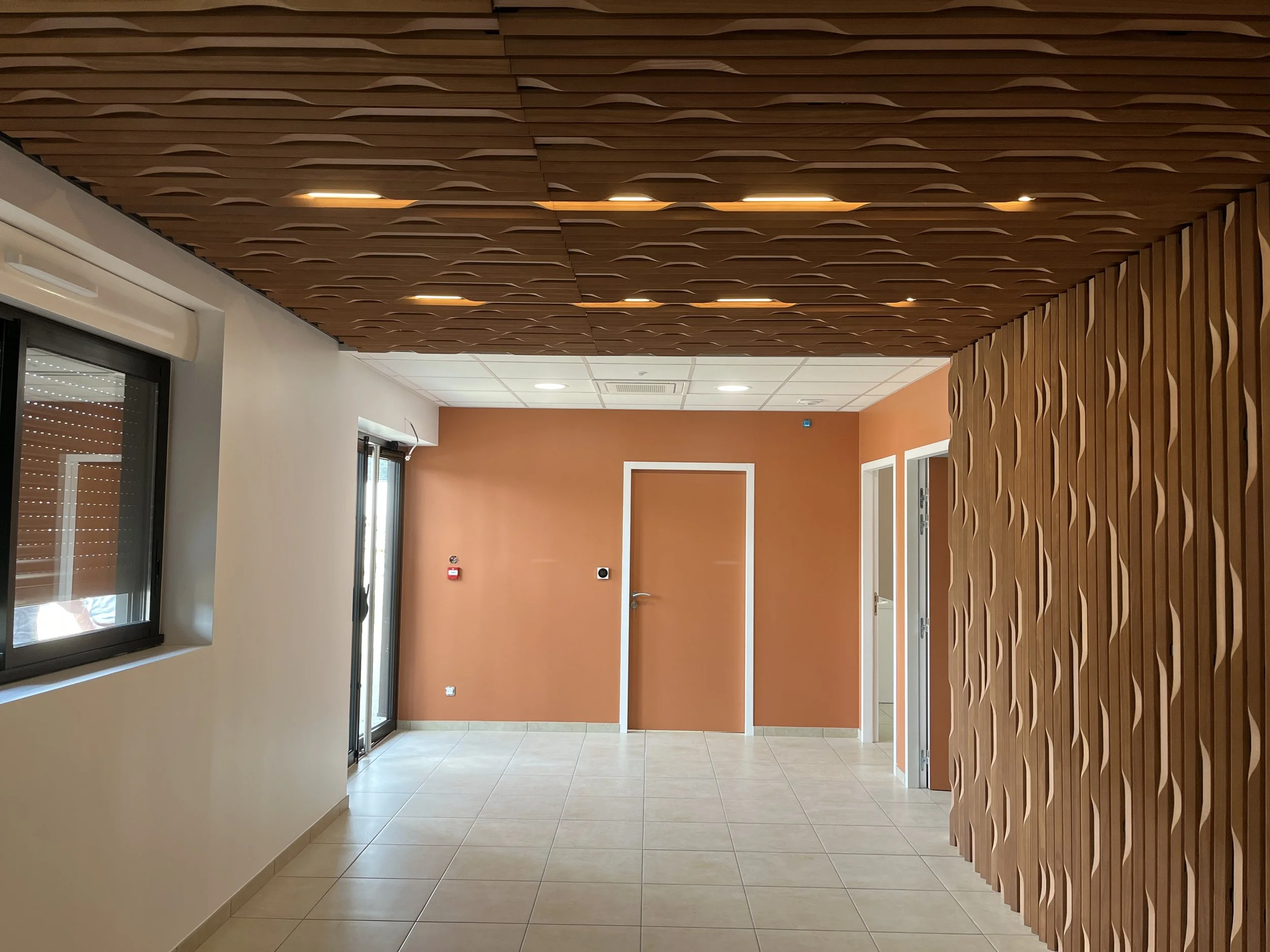 Intérieur d'un bâtiment moderne avec murs orange, plafond en bois avec des motifs sculptés, carrelage beige au sol, fenêtres avec volets roulants, et une porte blanche.