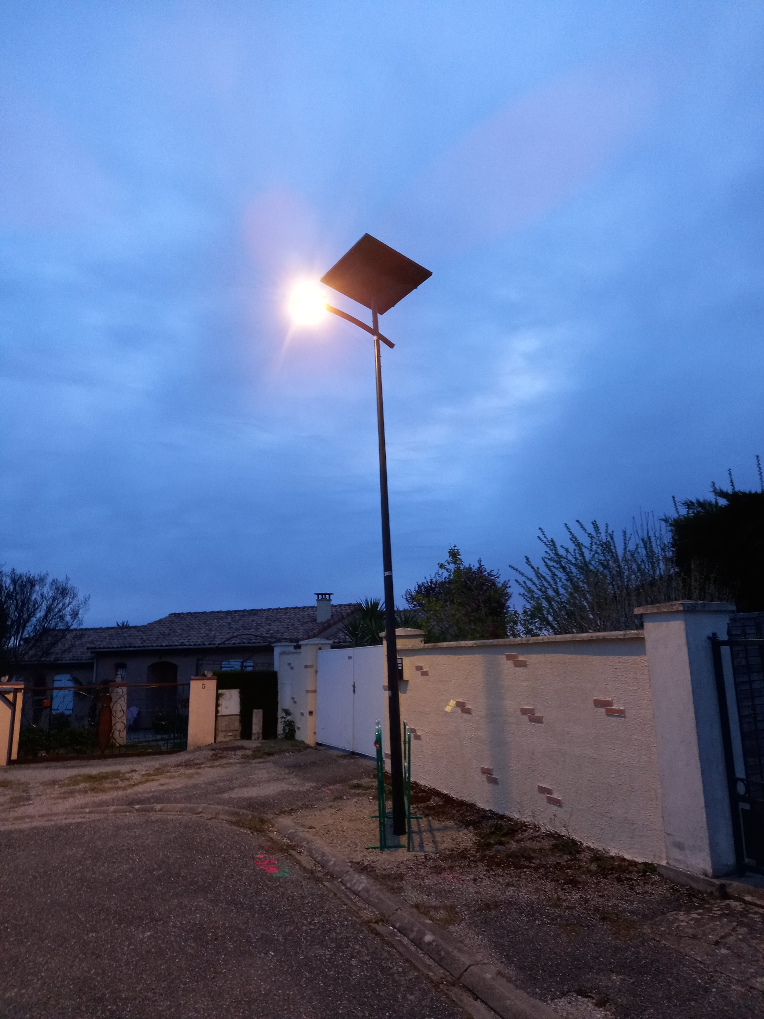 Un lampadaire avec un panneau solaire fixé en haut, éclairant une rue résidentielle en soirée ou à l'aube.