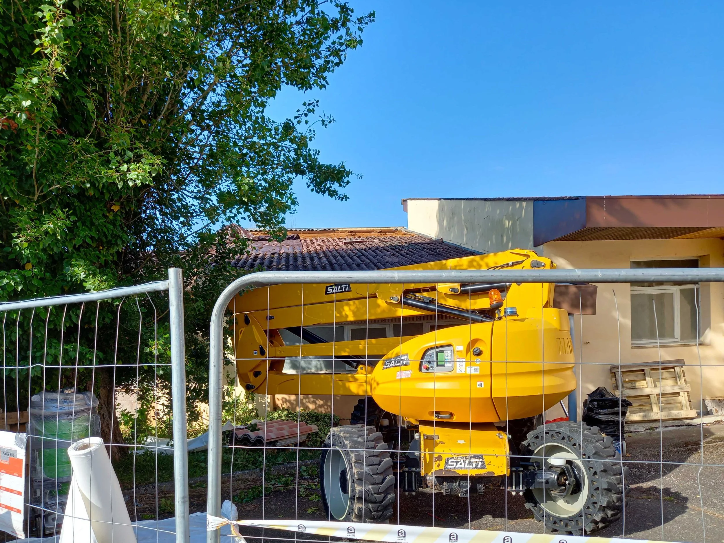 Une mini pelle jaune sur un chantier à l'extérieur, derrière une barrière métallique, avec des bâtiments et un arbre à proximité.