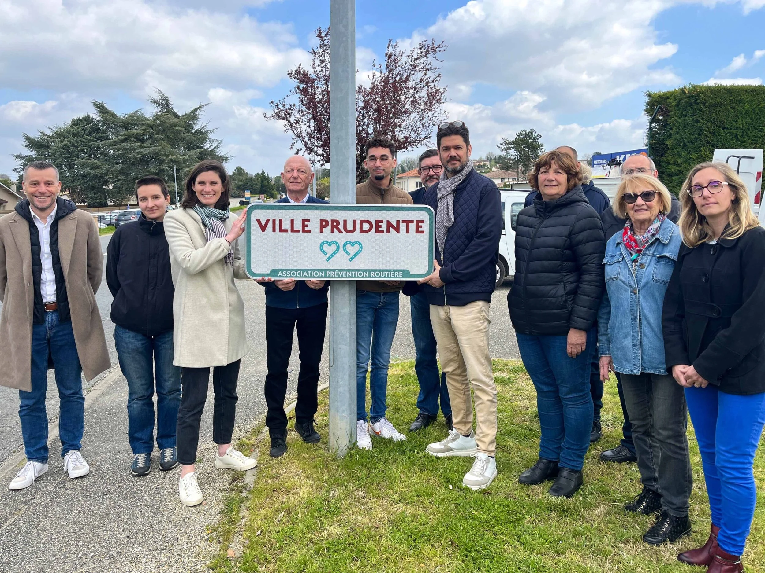 Groupe de personnes posant autour d'un panneau de signalisation avec le texte "Ville Prudente" et un symbole de coeurs, lors d'une réunion ou d'une cérémonie en plein air.