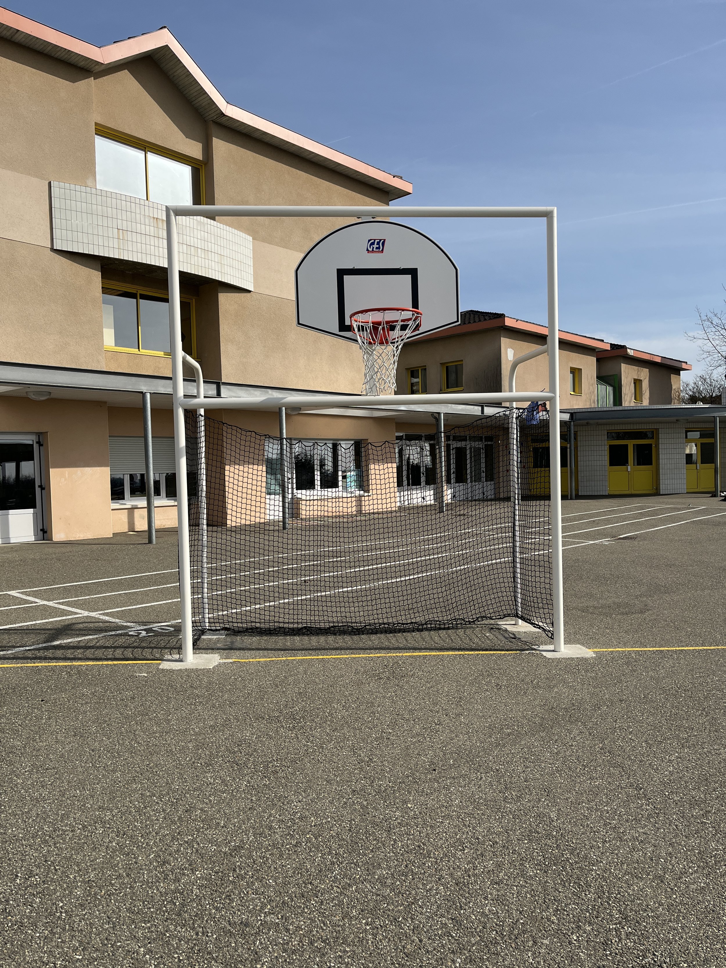 Un terrain de sport avec un panier de basketball et un goal pour le football, situé dans une cour d'école ou un centre éducatif, avec un bâtiment en arrière-plan et un ciel clair.