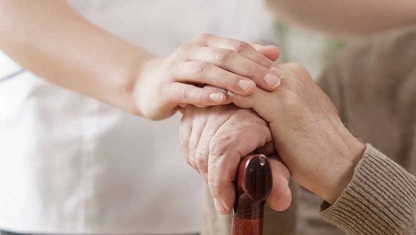 Une personne âgée tenant une canne pour se soutenir, tandis qu'une autre personne lui tient la main en signe de soutien ou de compassion.