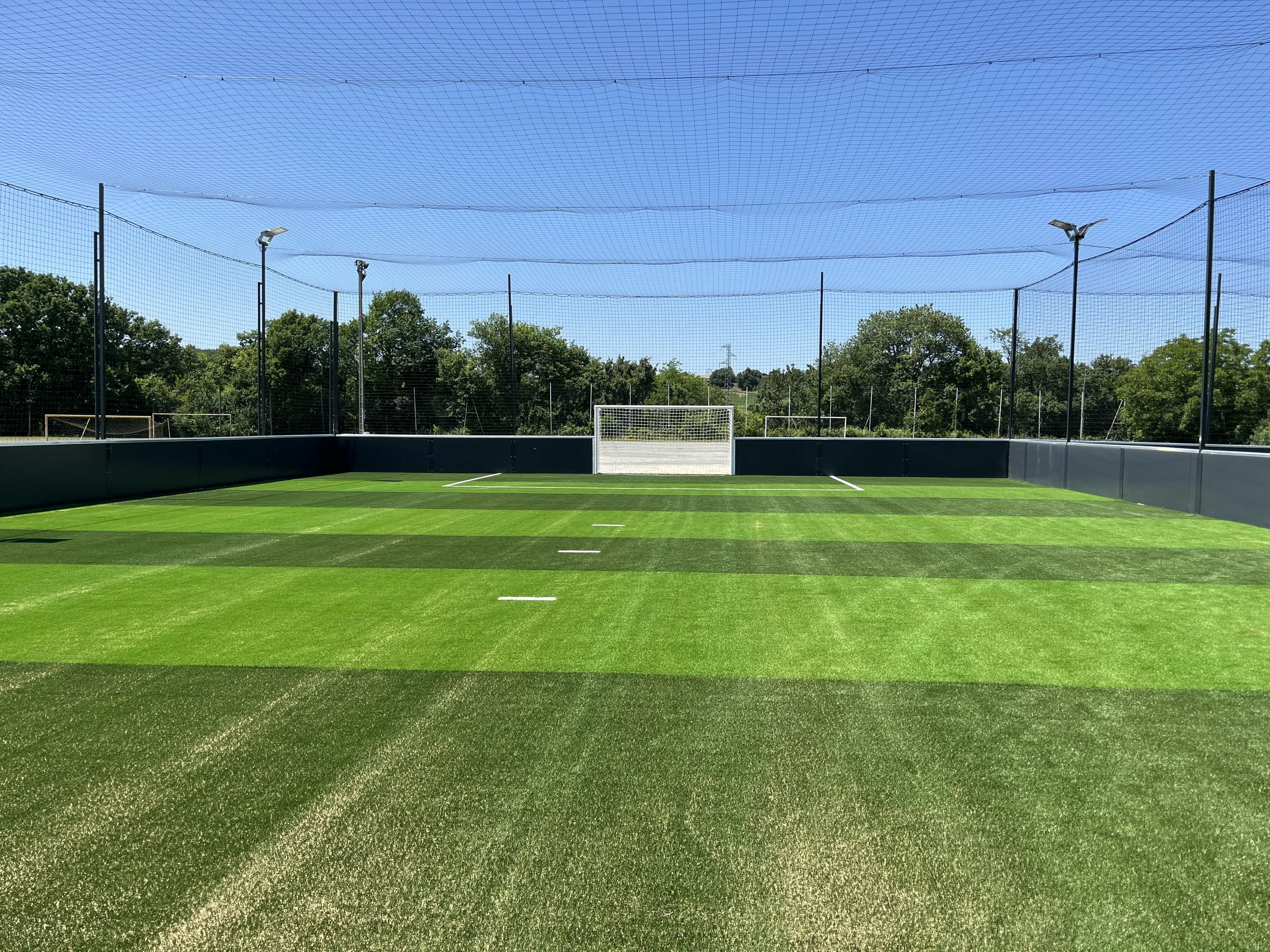 Terrains de football en gazon synthétique avec but en arrière-plan, entourés de filets et d'éclairage, sous un ciel bleu.