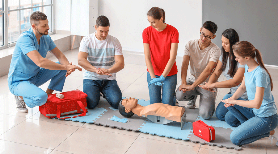 Groupe de jeunes étudiants en formation aux premiers secours, pratiquant la réanimation sur un mannequin en salle. Présence d'une trousse de secours et de matelas de formation.