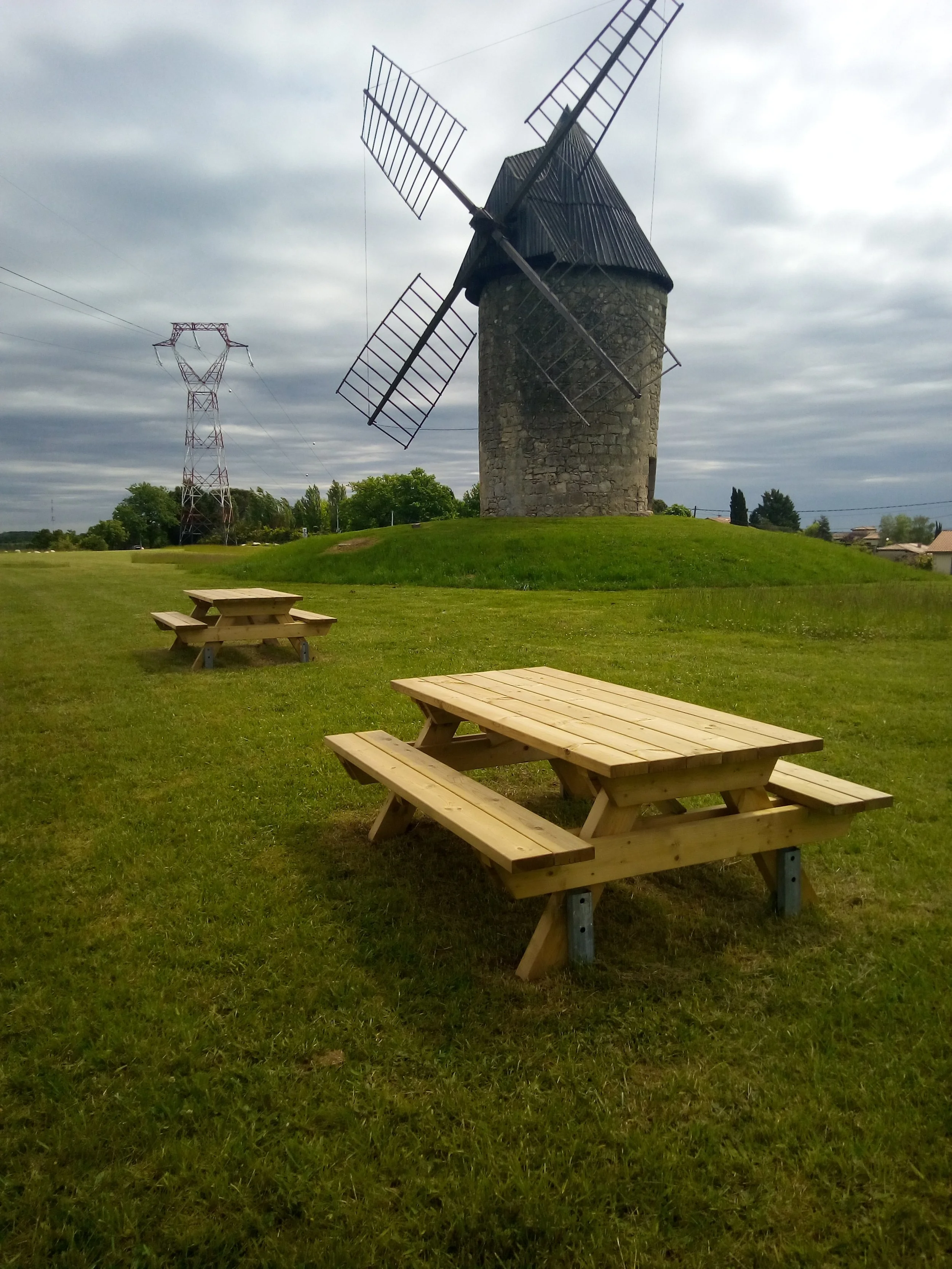 Un moulin à vent en pierre avec des ailes en bois, situé sur une colline, avec des tables de pique-nique en bois dans l'herbe verte environnante, sous un ciel nuageux.