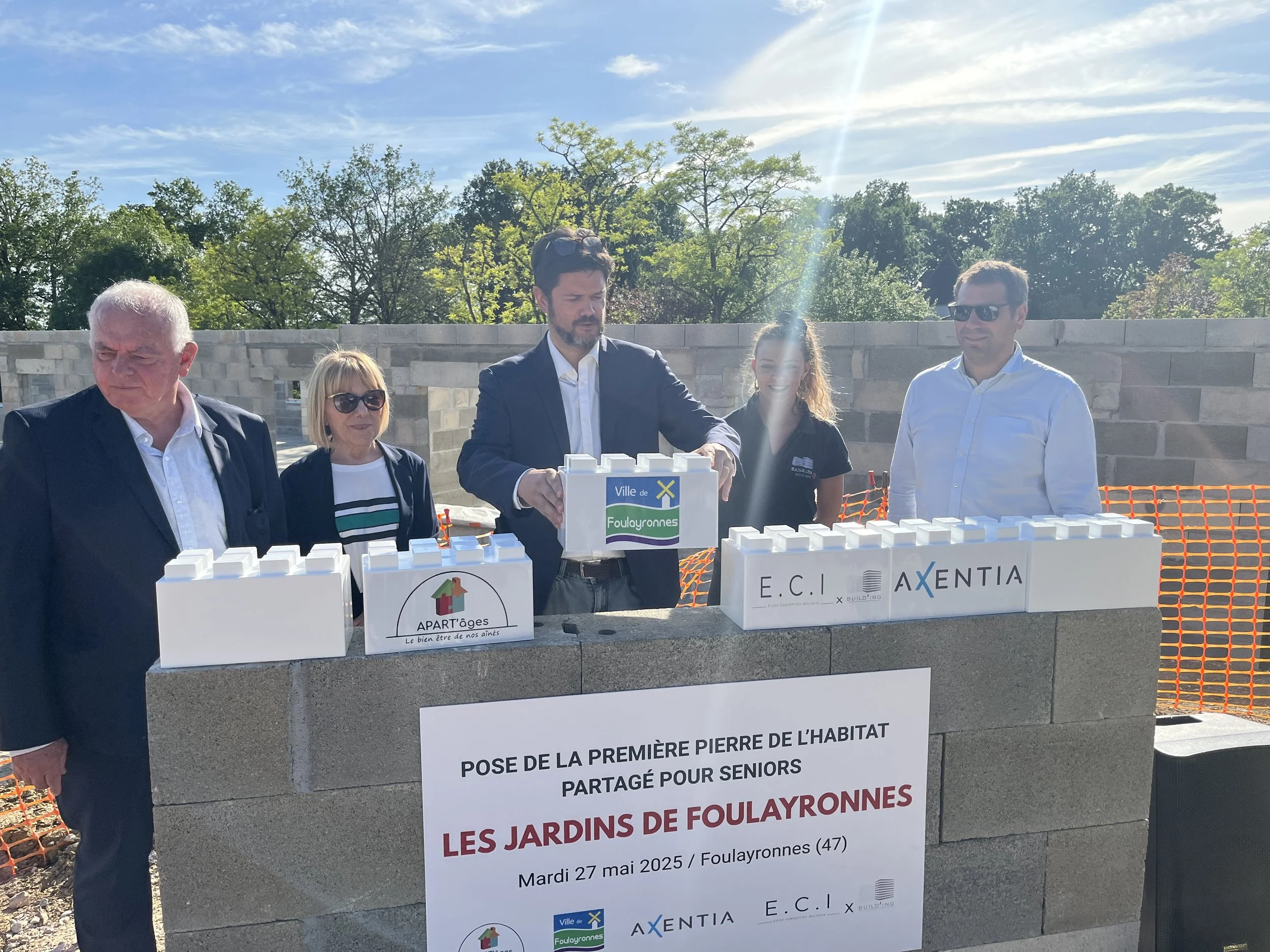 Cinq personnes se tiennent derrière un mur de béton lors d'une cérémonie. Elles participent à la pose de la première pierre d'un projet partagé pour seniors dans les jardins de Foulayronnes, le 27 mai 2025. Le soleil brille et le ciel est clair, avec quelques nuages. Certaines personnes portent des lunettes de soleil.