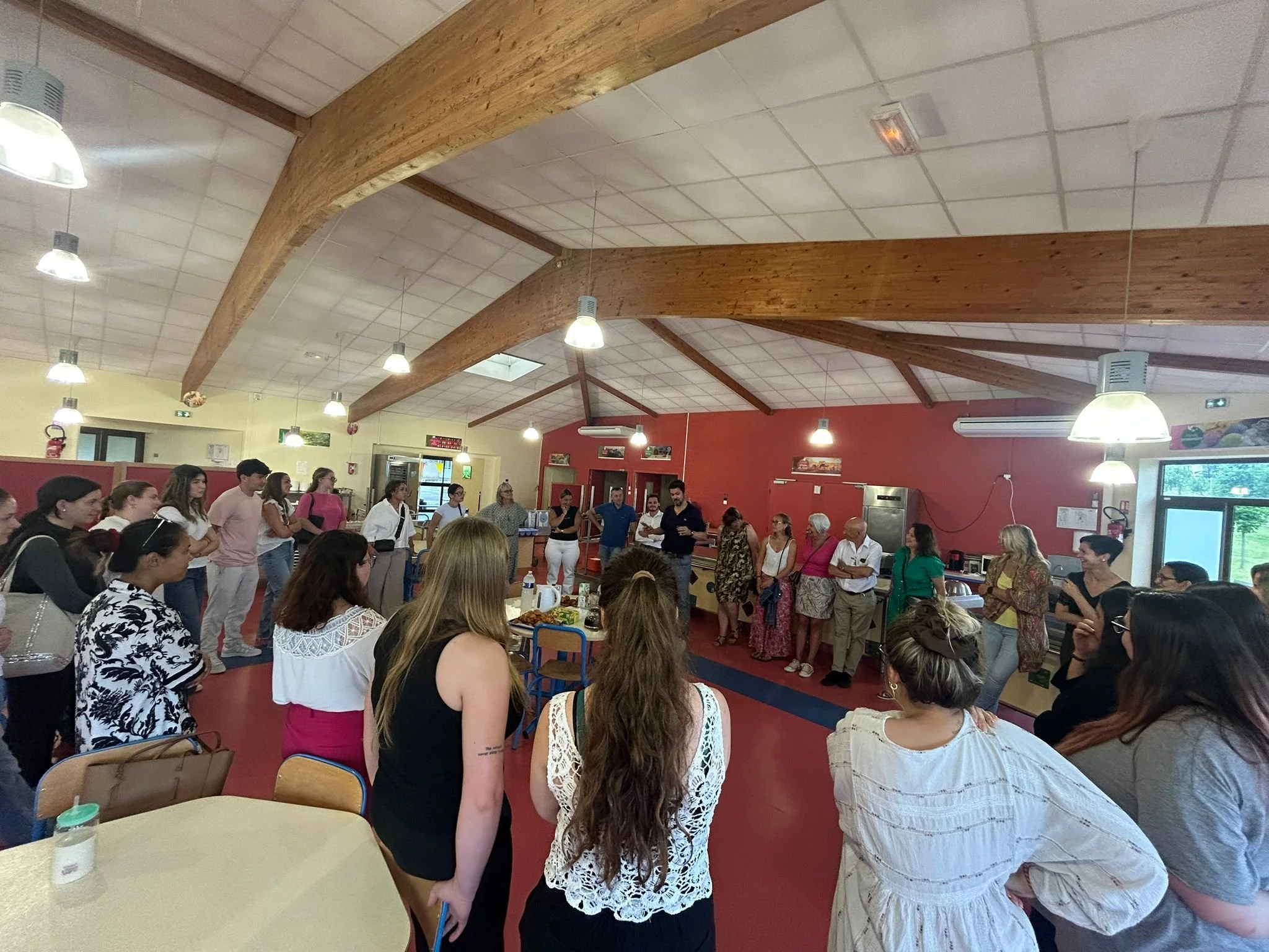 Groupe de personnes rassemblées dans une salle, probablement pour une réunion ou un événement social, avec des tables et un plafond en bois.