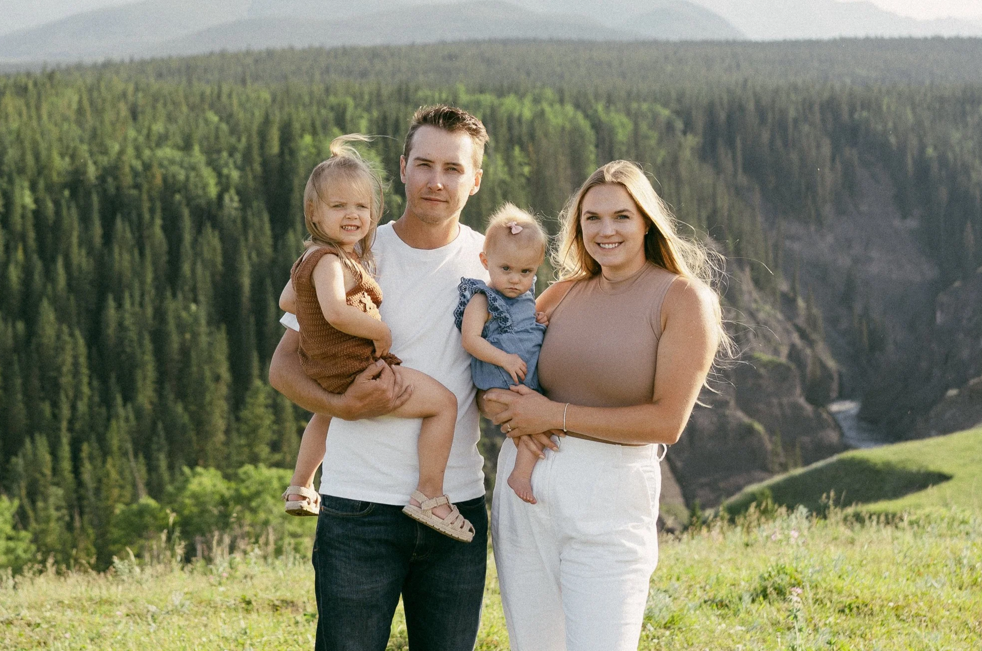 A happy family of four in a scenic outdoor setting with green hills and a forested canyon in the background. The father holds a young girl in a brown dress, while the mother holds a baby girl in a blue outfit.