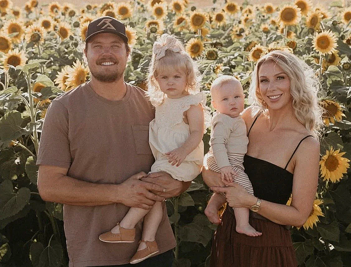 A happy family of four posing among sunflowers outdoors during daytime. The father and mother, each holding a child, stand smiling at the camera.