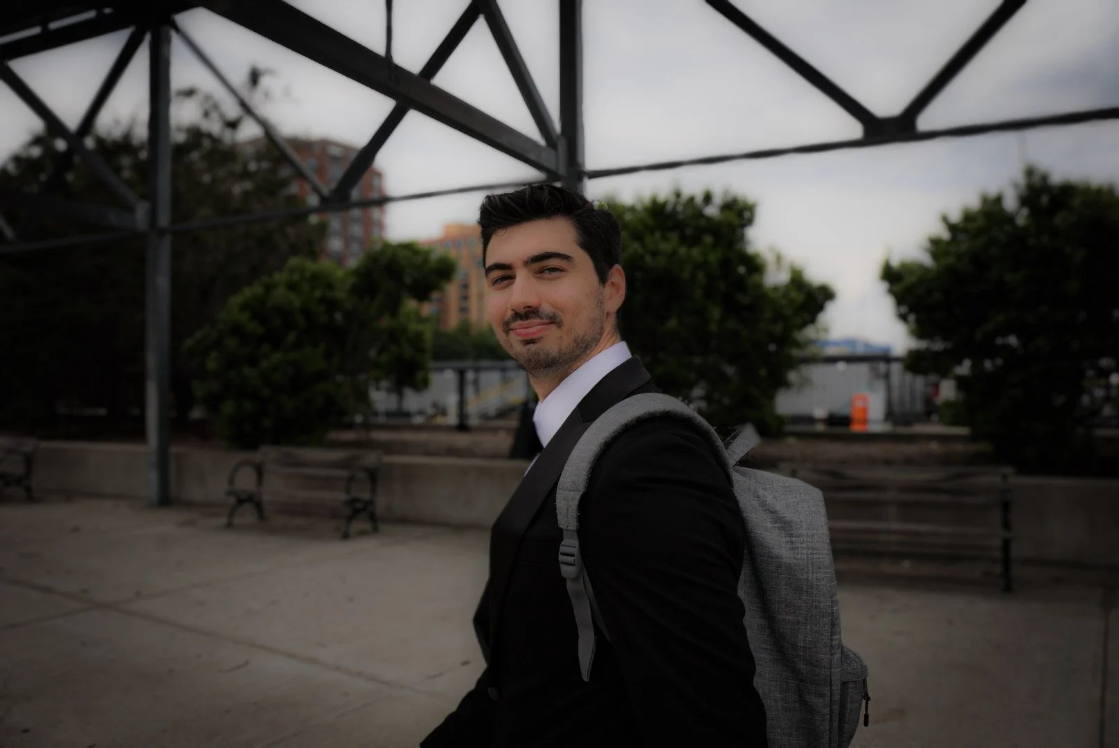 A young man with dark hair and a beard, wearing a black suit and white shirt, carrying a gray backpack, standing outdoors under a metal structure with trees and buildings in the background.