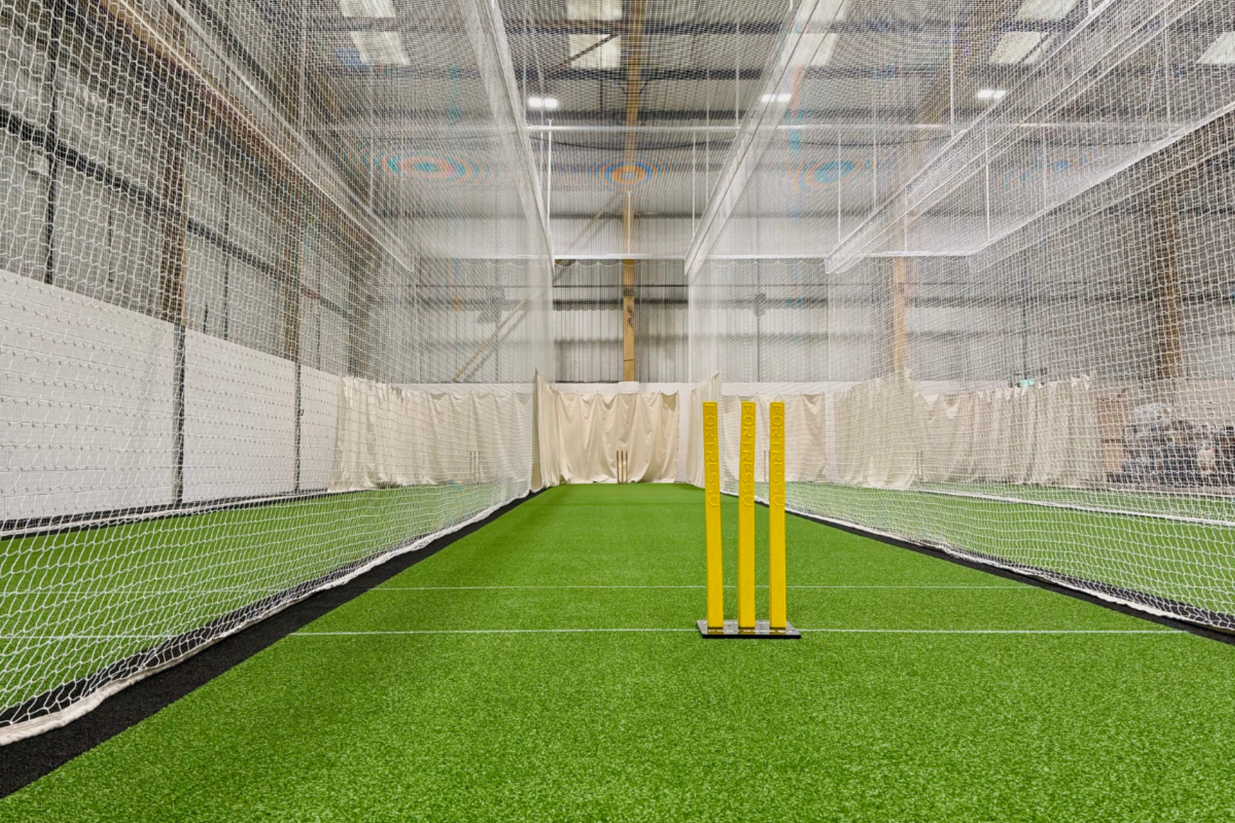 Indoor cricket net with green artificial turf, three yellow wickets, and protective netting surrounding the playing area.