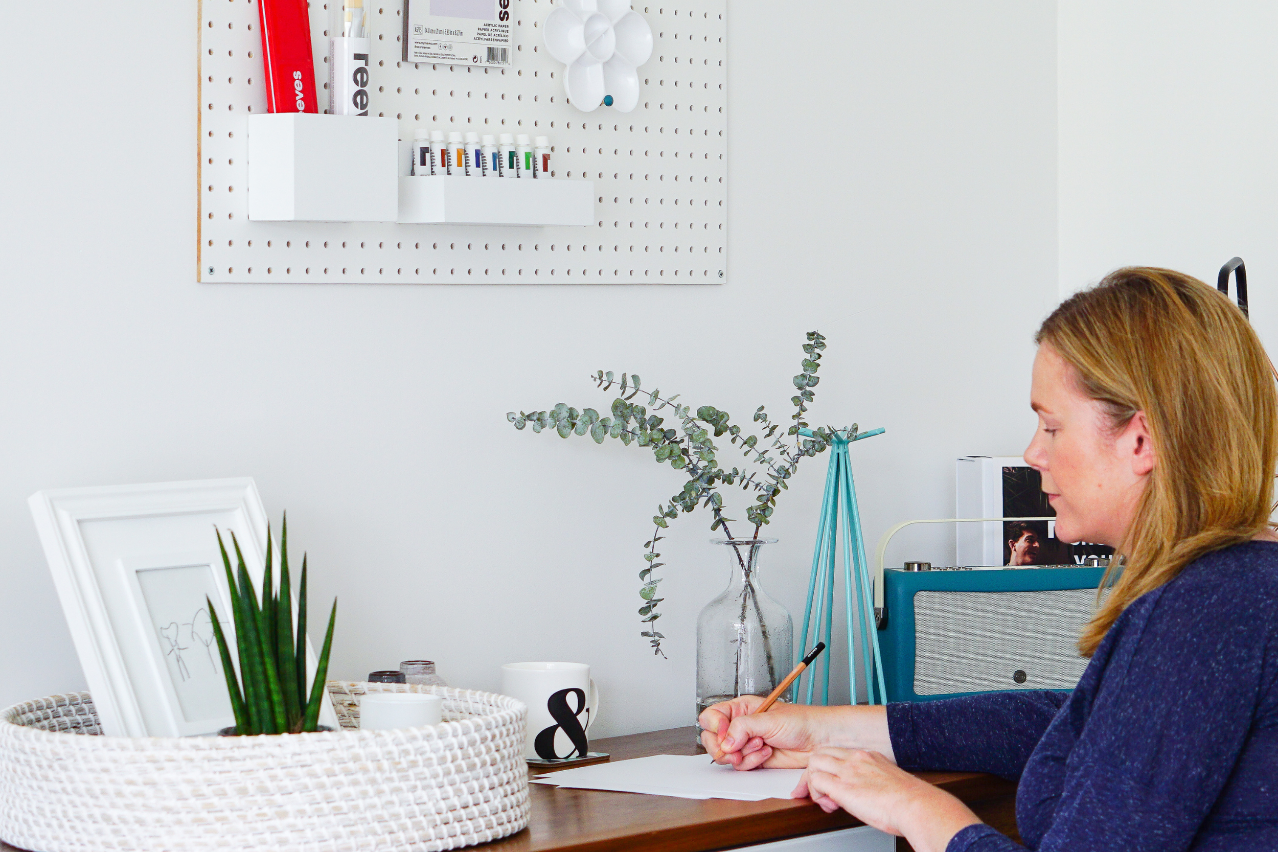 A woman with reddish-brown hair sitting at a desk drawing on paper with a pencil. The desk has a white basket with a framed sketch and a green plant, and a large glass vase with eucalyptus leaves. The background has a white wall with a pegboard holding papers, markers, and other small items.