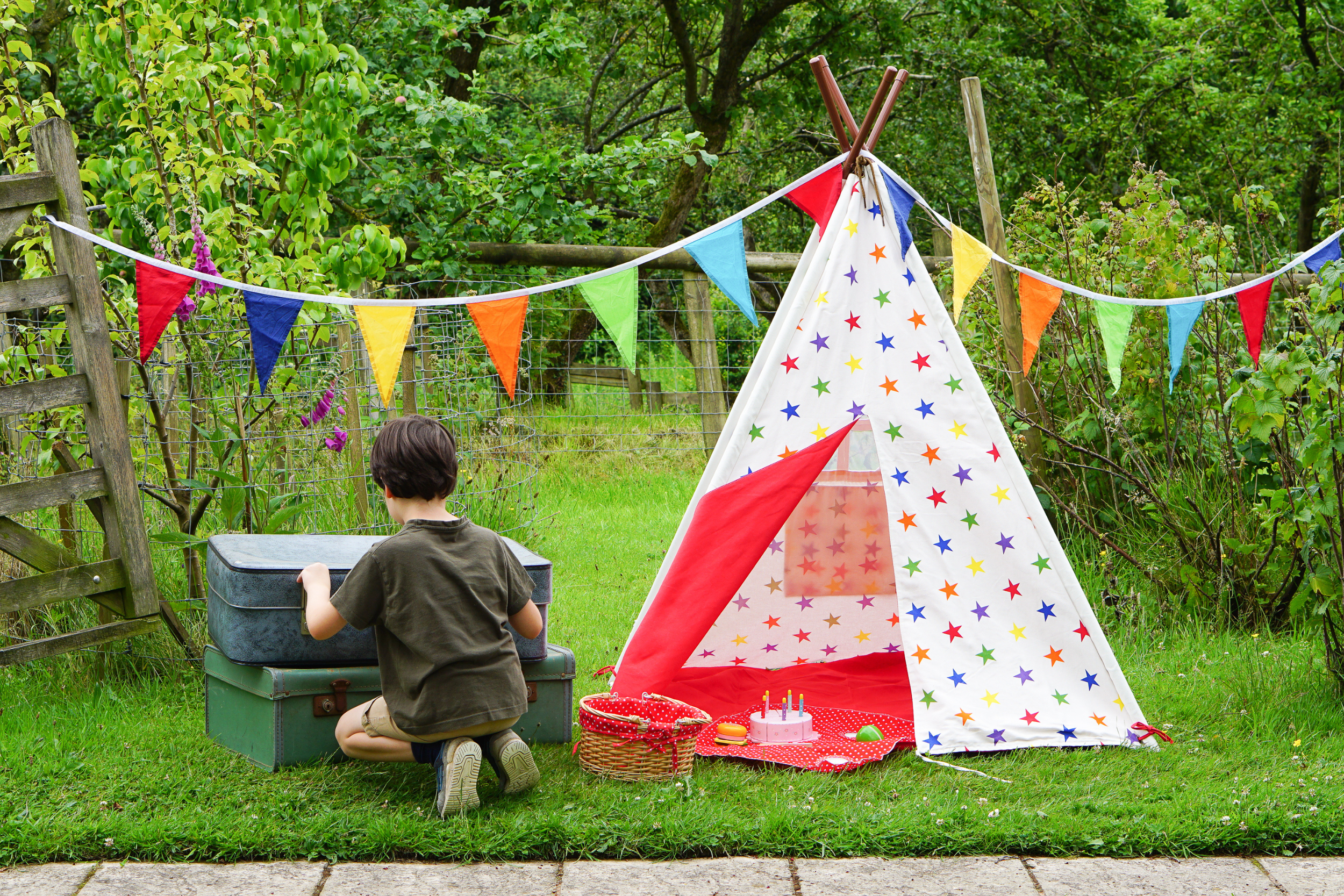 A young boy kneeling next to an open suitcase outside, near a colorful unicorn-themed play tent decorated with stars, with a birthday cake and party favors in front, and festive bunting hanging overhead in a lush green garden.