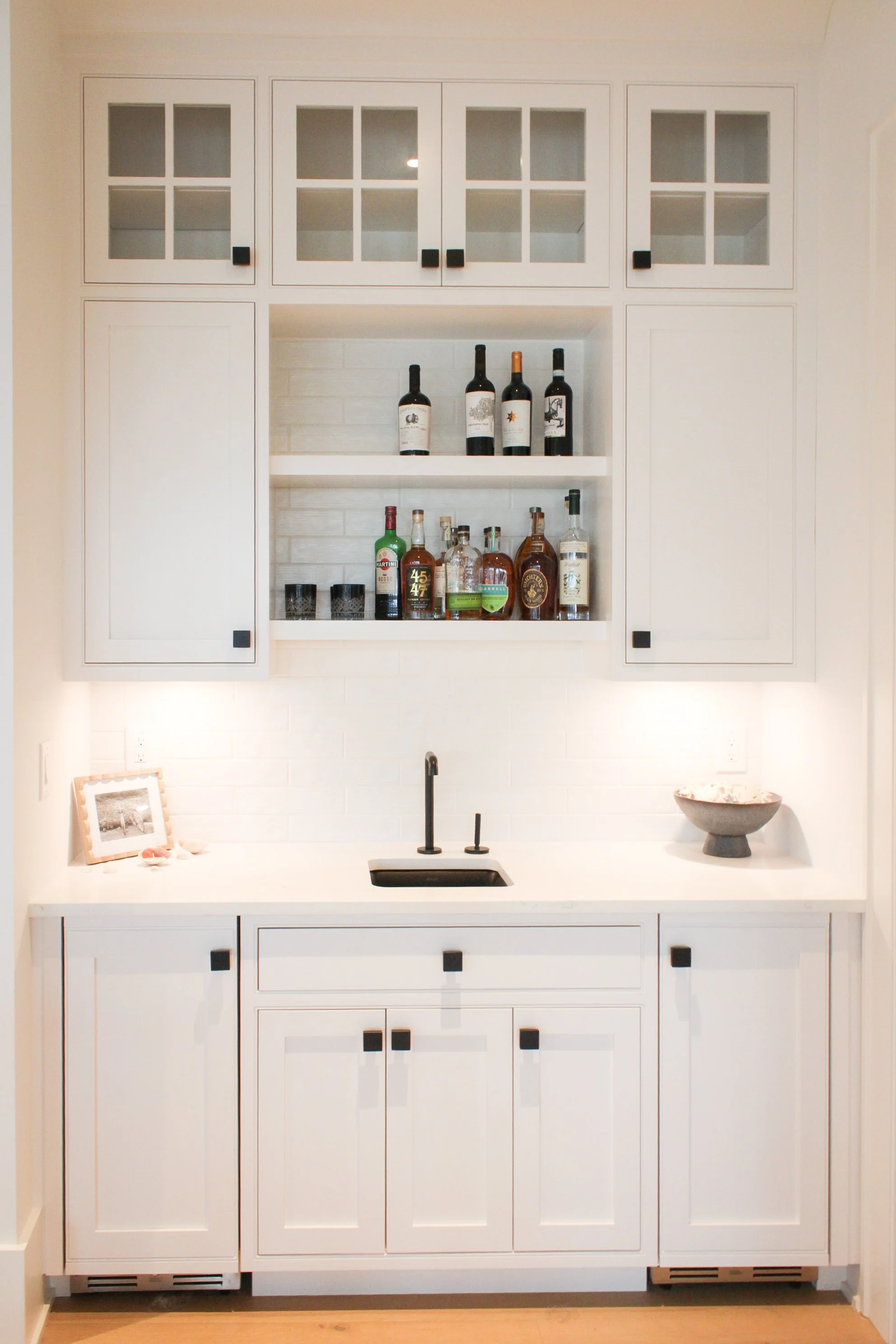 White kitchen cabinet with black square knobs, open shelves with liquor bottles, small sink, black faucet, and a bowl on the countertop.