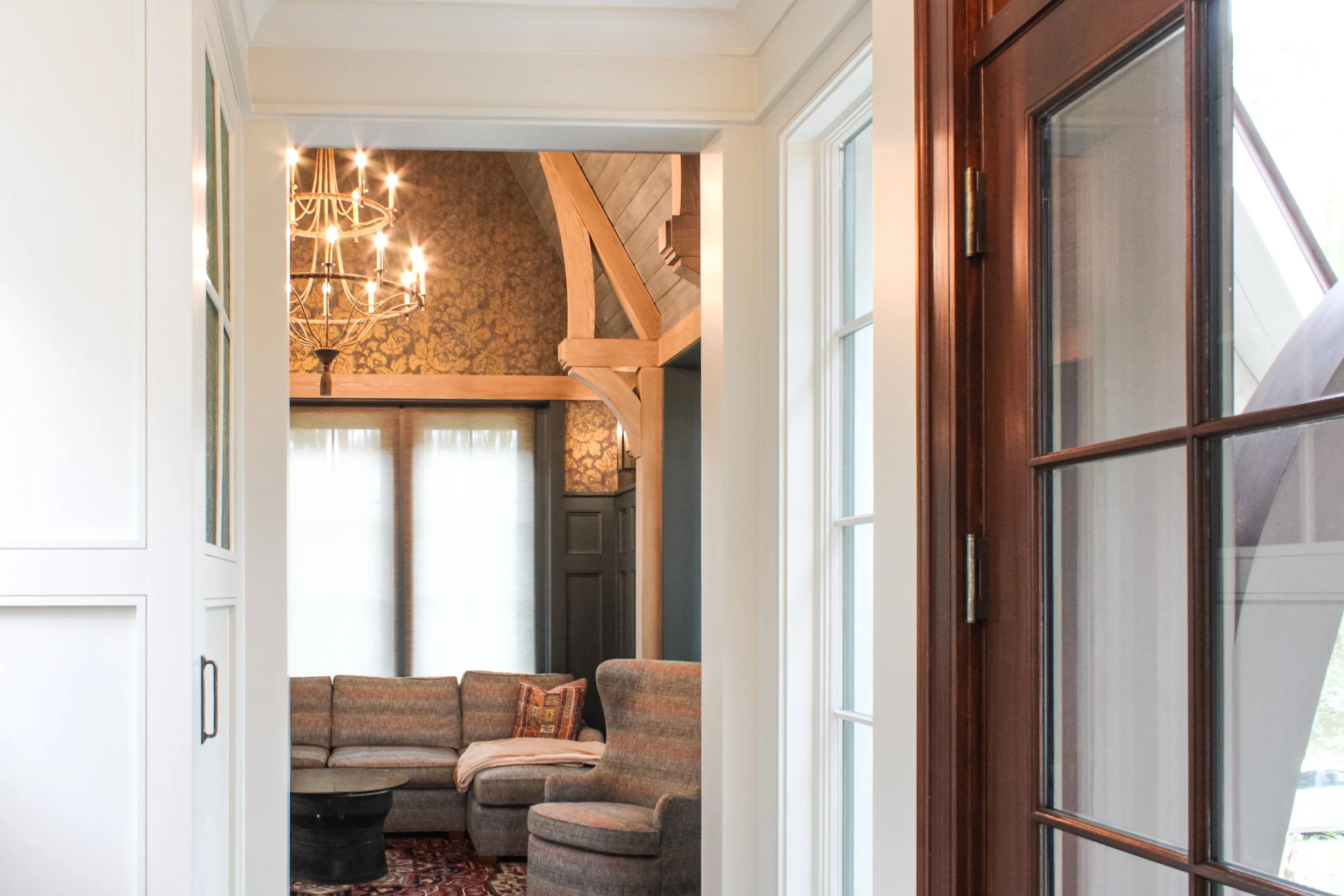 Interior view of a living room with a brown sectional sofa, a round coffee table, and a window with white curtains. A chandelier hangs from the ceiling, and wooden beams are visible near the ceiling. Part of a glass-paneled door is in the foreground on the right.