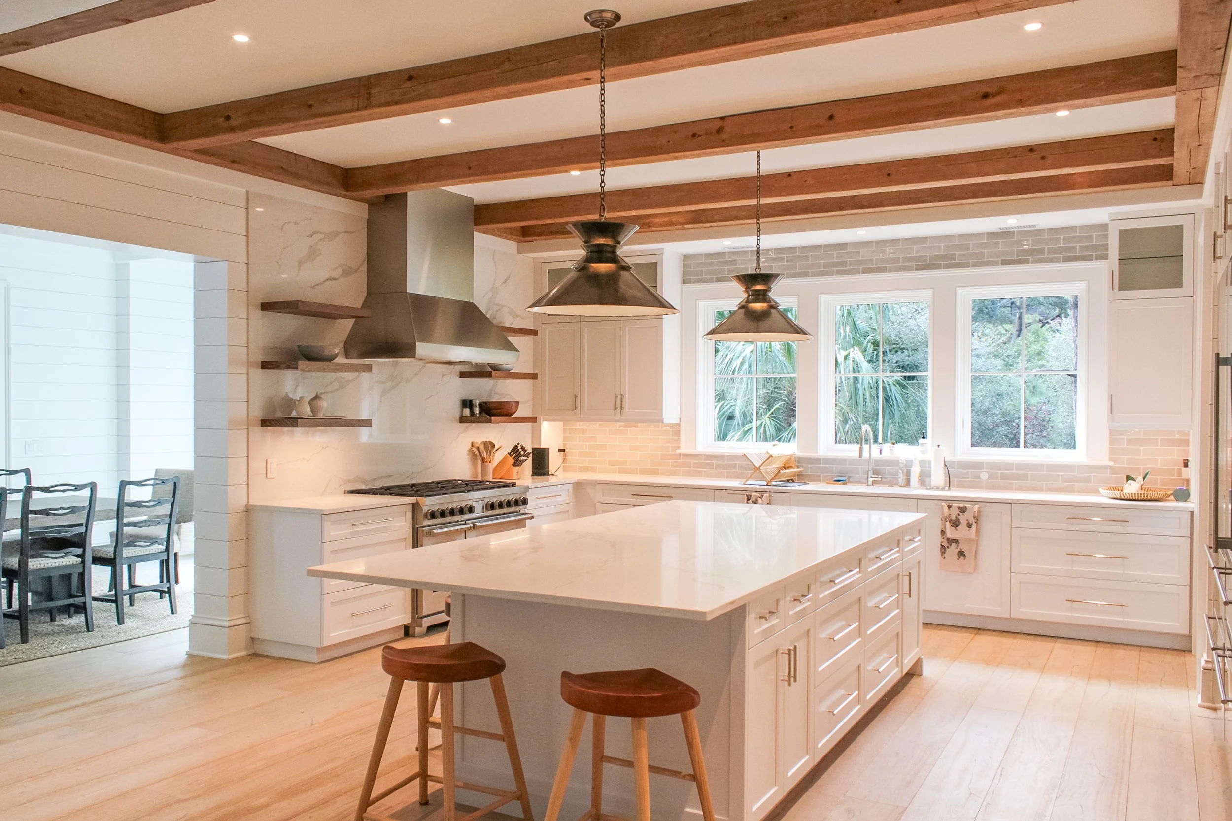 Bright kitchen with white cabinets, marble backsplash, wooden ceiling beams, large window view of greenery, central island with stools, stainless steel appliances, and pendant lights.