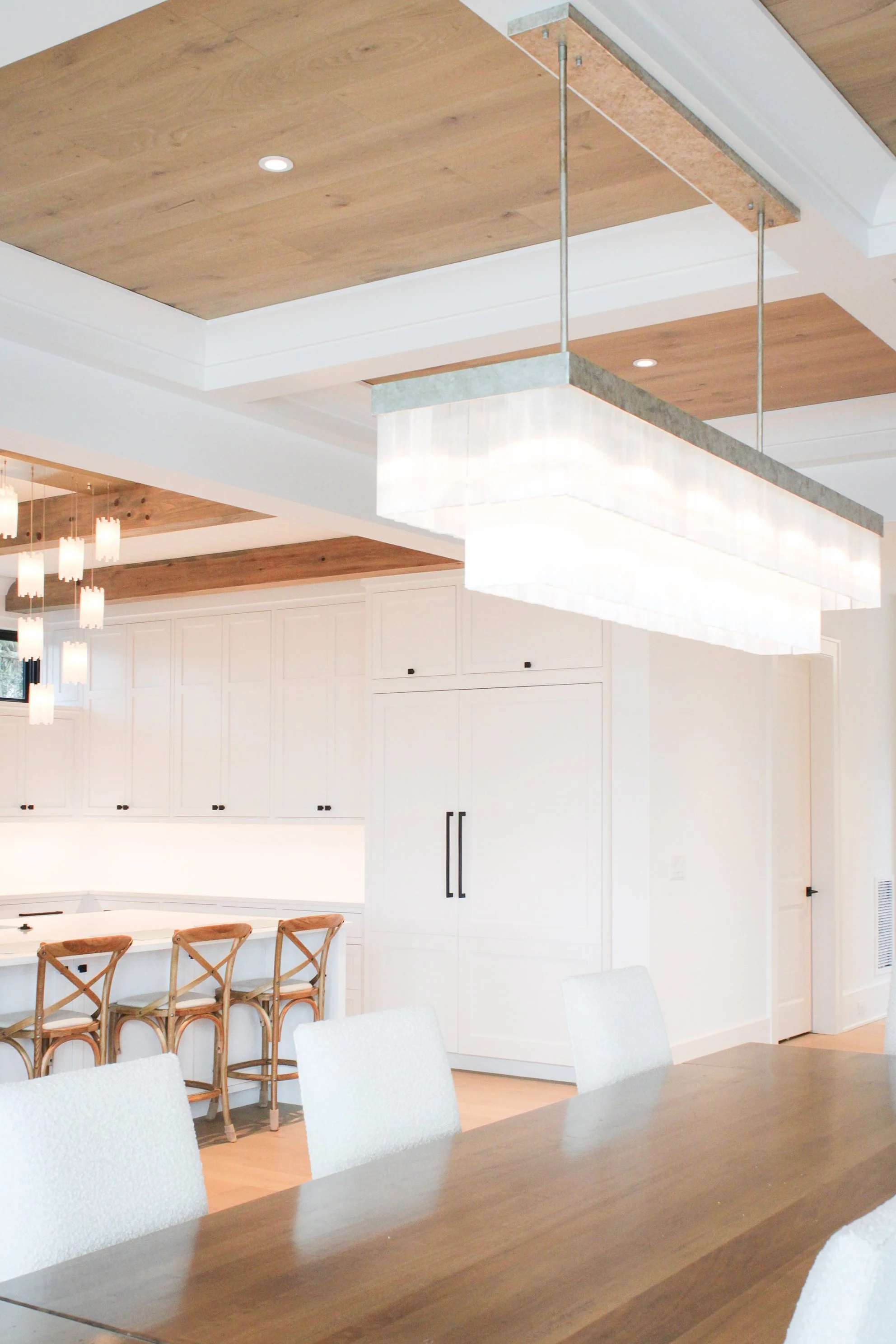 Modern kitchen and dining area with a large rectangular hanging light fixture, wooden ceiling panels, white cabinetry, and light-colored chairs around a wooden table.