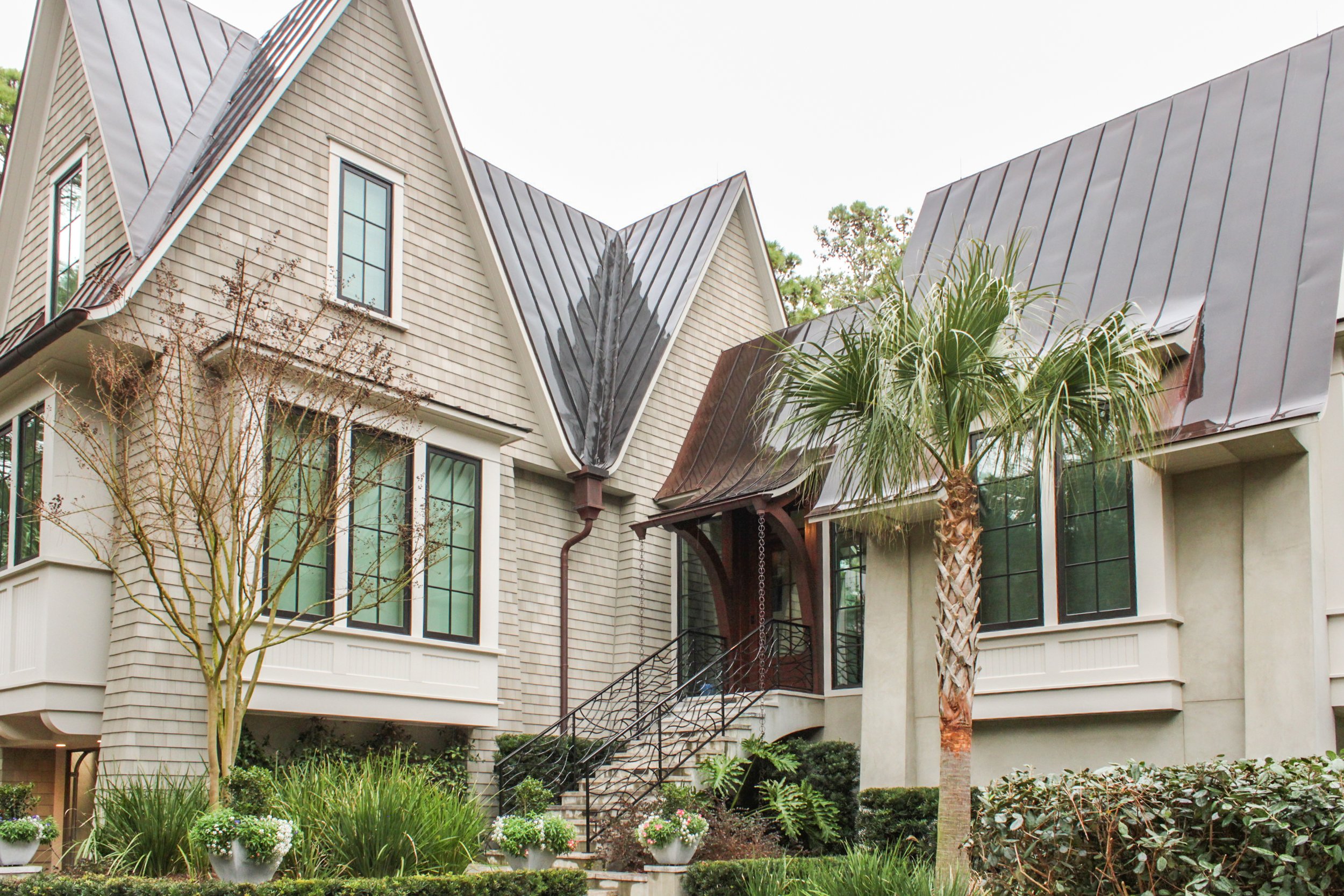 Exterior of a modern house with angular metal roofs, large windows, landscaped garden with small trees, shrubs, flowers, and a palm tree.