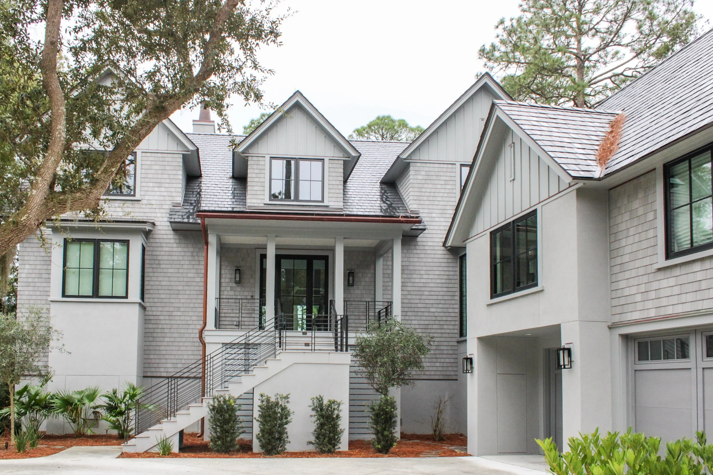 Front view of a modern two-story house with stairs leading to the front door, surrounded by trees and landscaped yard.