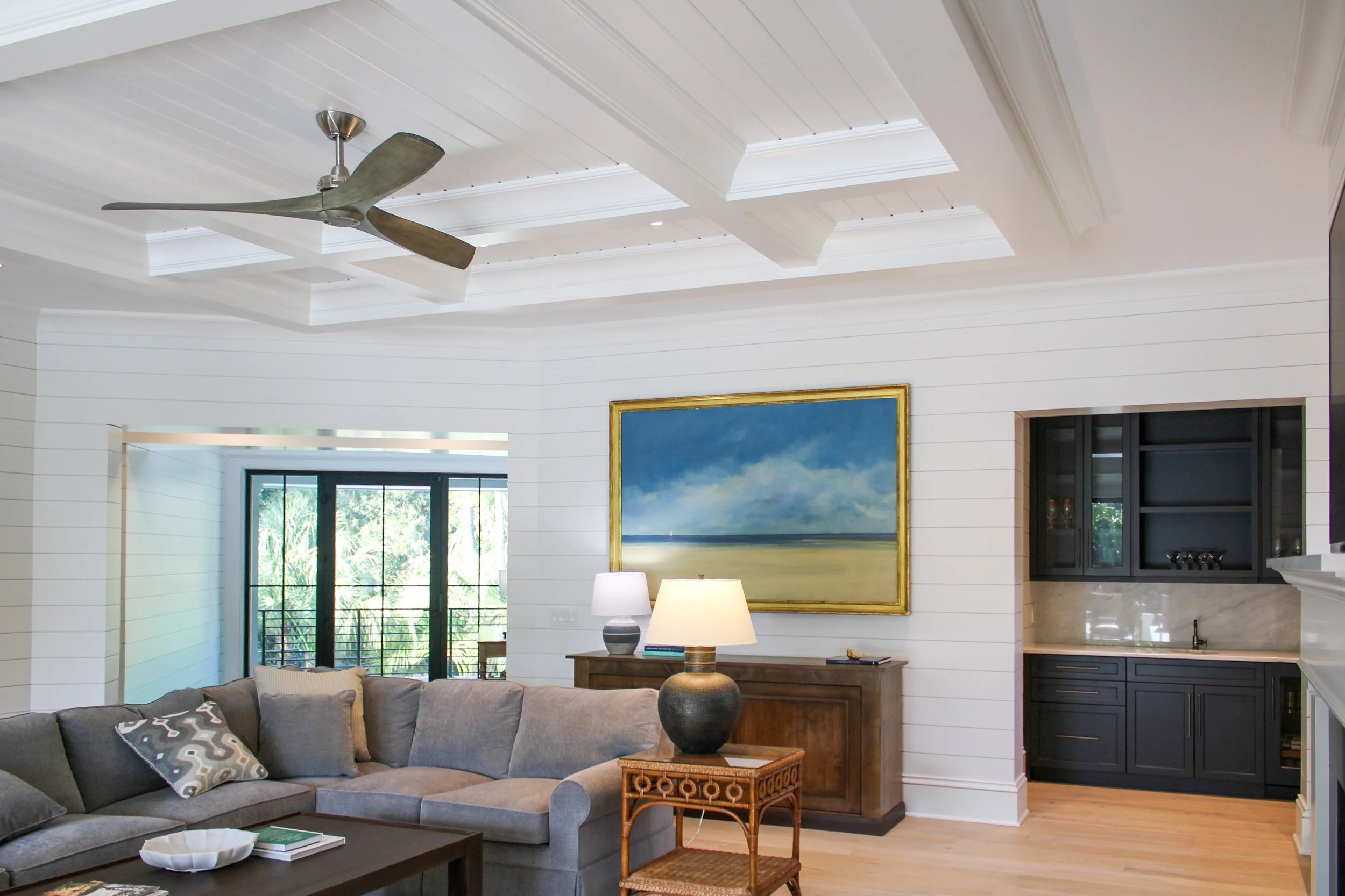 Living room with white shiplap walls, a ceiling fan, an abstract landscape painting, table lamps, a gray sectional sofa with patterned pillows, and a black kitchen with marble countertops visible in the background.
