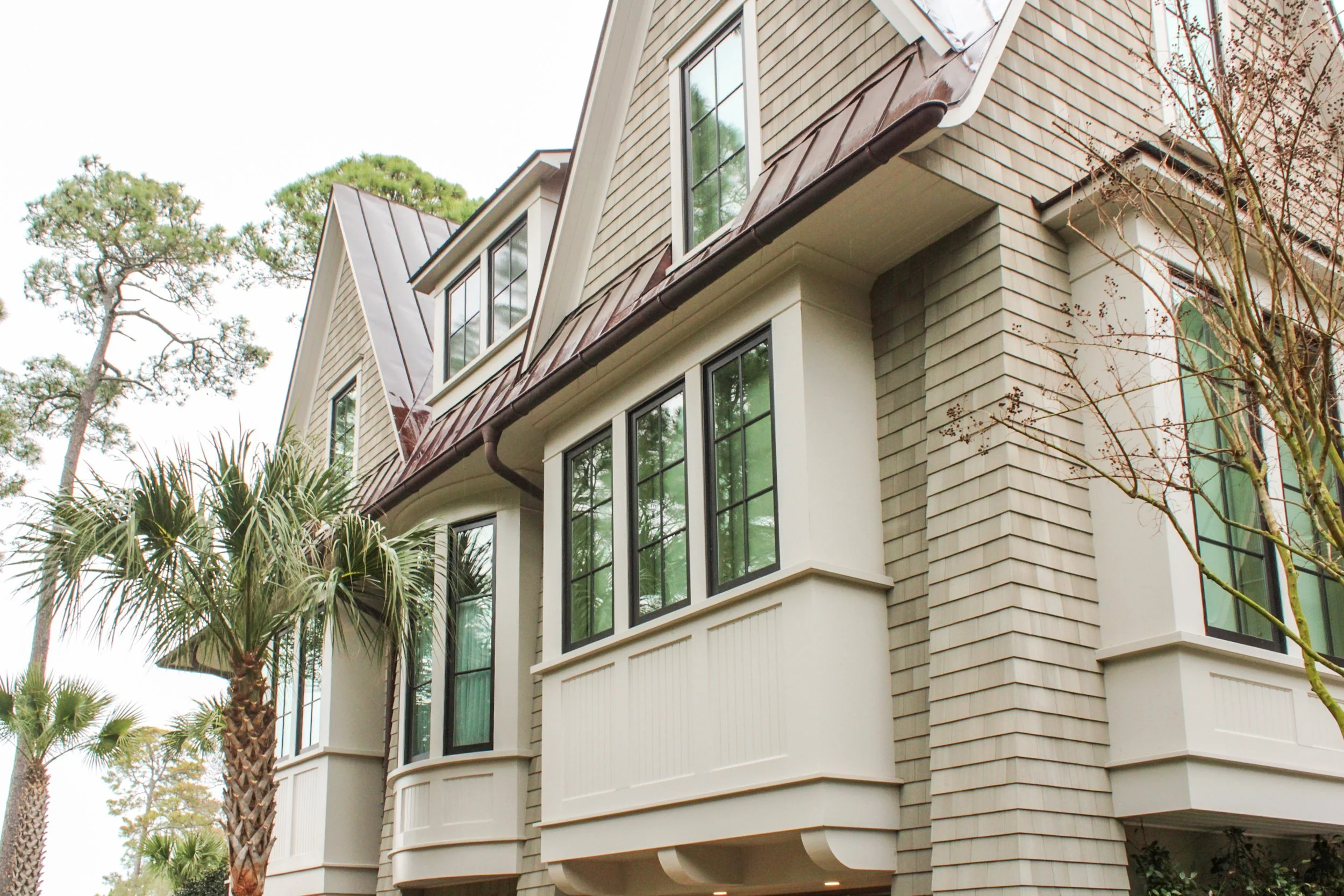 Close-up of a modern multi-story house with beige siding, dark brown roof, and large windows, surrounded by trees and palm trees.