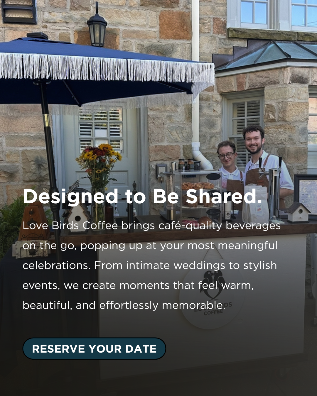 Two men smiling behind a coffee stand with flowers, jars, and coffee equipment outside a stone building with windows and a navy blue umbrella.