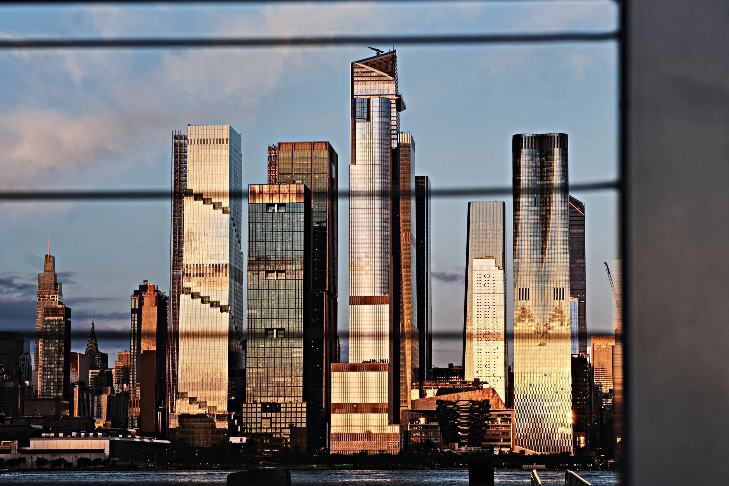 View of New York City skyline with tall glass skyscrapers reflecting sunlight, seen through window blinds.