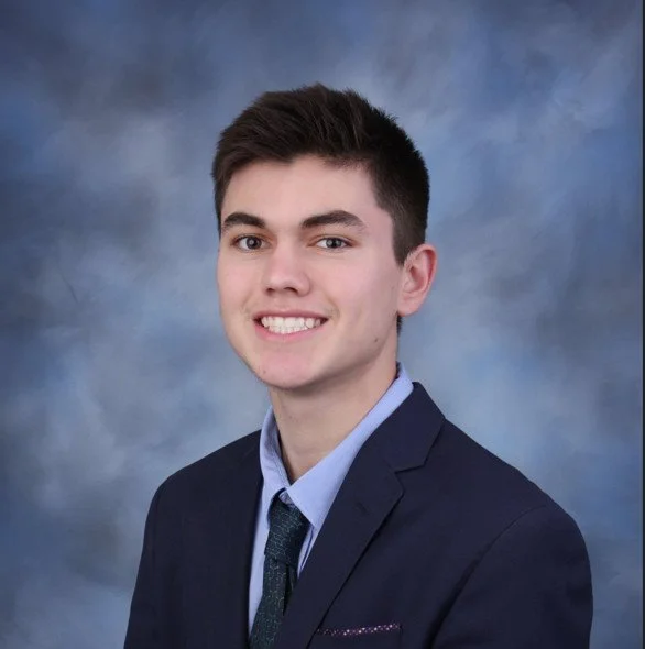 Young man in a navy suit, light blue shirt, and patterned tie, smiling against a blue and gray cloudy background.