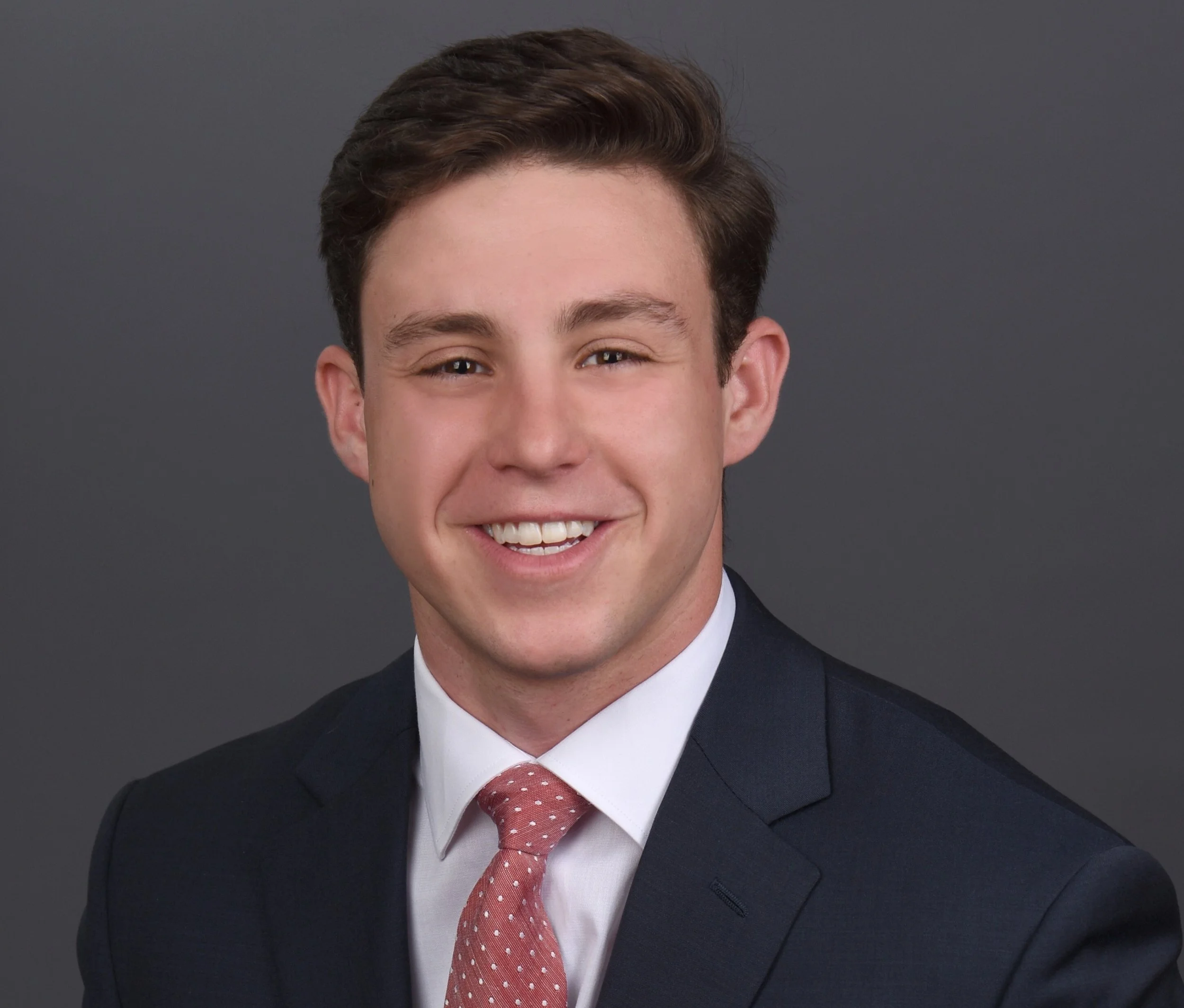 Close-up of a young man smiling, wearing a dark suit, white shirt, and a red tie with white polka dots, against a dark gray background.