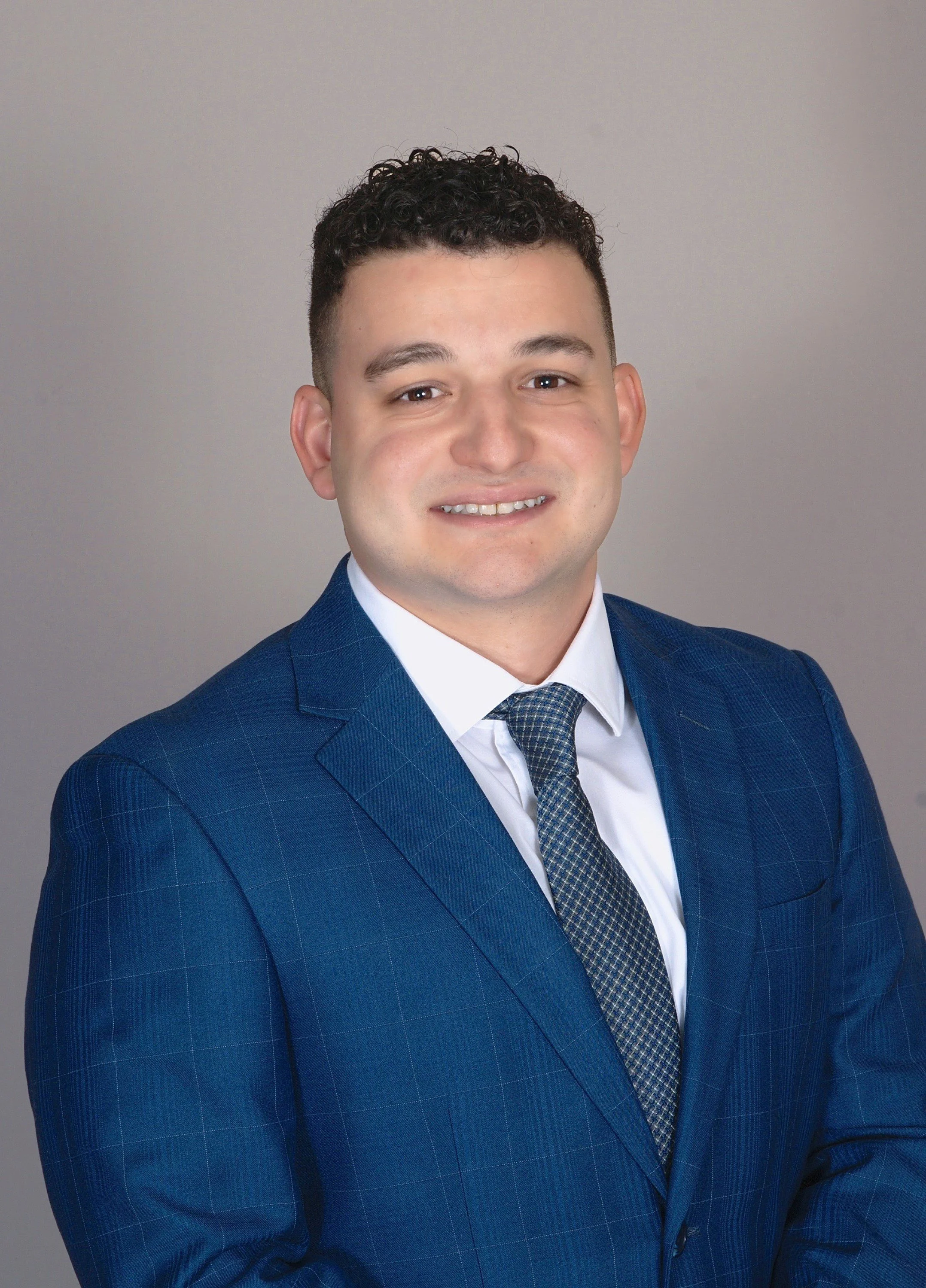 Professional headshot of a man in a blue plaid suit, white shirt, and patterned tie, smiling against a plain grey background.