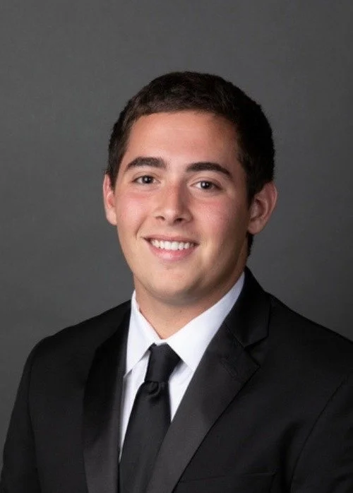 A young man wearing a black suit and tie, smiling against a gray background.