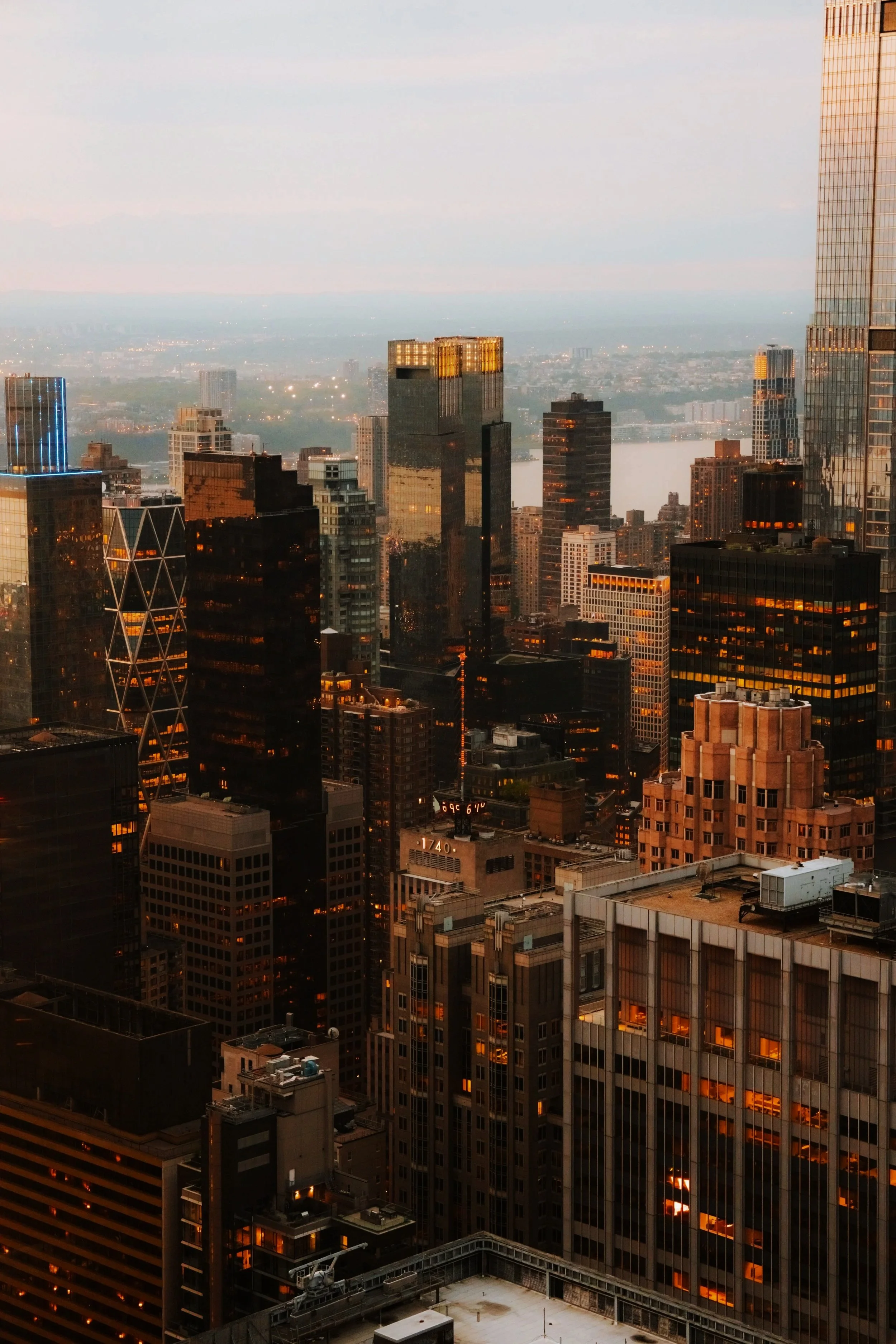 A city skyline at dusk featuring tall skyscrapers with illuminated windows, some with reflective glass facades, and a river in the background. Buildings are lit with warm and cool tones, with some having LED lighting accents.