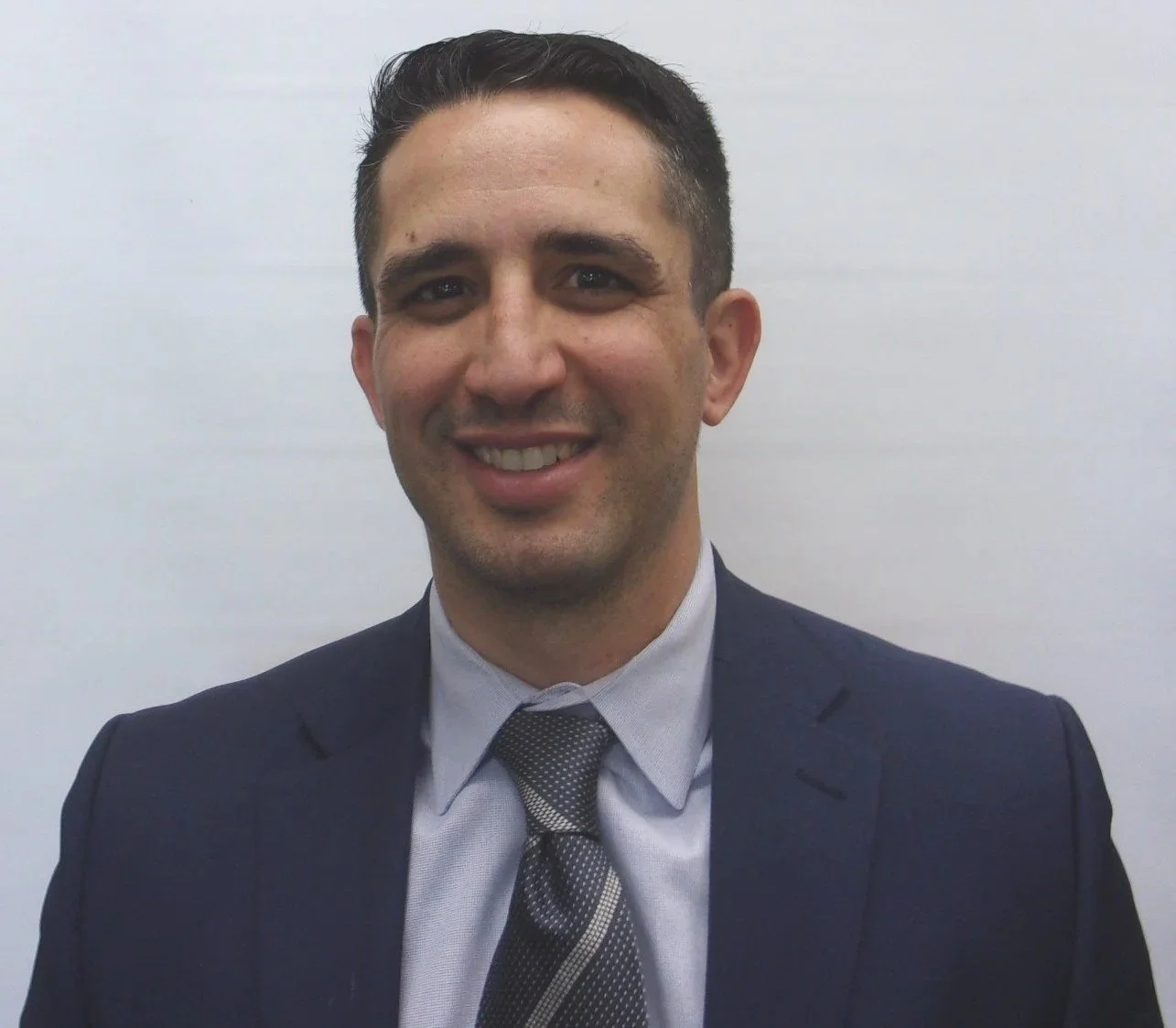 A smiling man dressed in a dark suit, light gray shirt, and patterned tie, standing in front of a plain light-colored background.