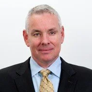 A portrait of a middle-aged man with short gray hair, wearing a dark suit, light blue dress shirt, and patterned tie, against a plain white background.