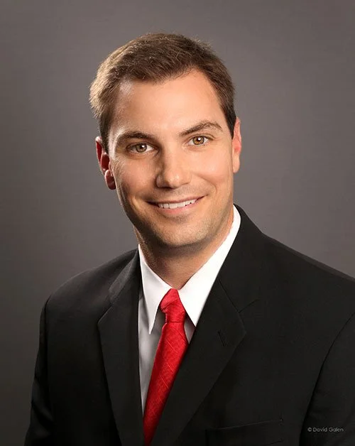 Professional portrait of a man in a black suit, white shirt, and red tie smiling against a gray background.