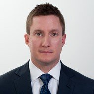 Headshot of a man with short brown hair wearing a navy suit, white shirt, and blue tie against a plain background.