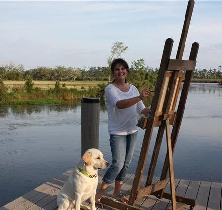 A woman painting on an easel near a body of water with a Labrador Retriever sitting nearby.