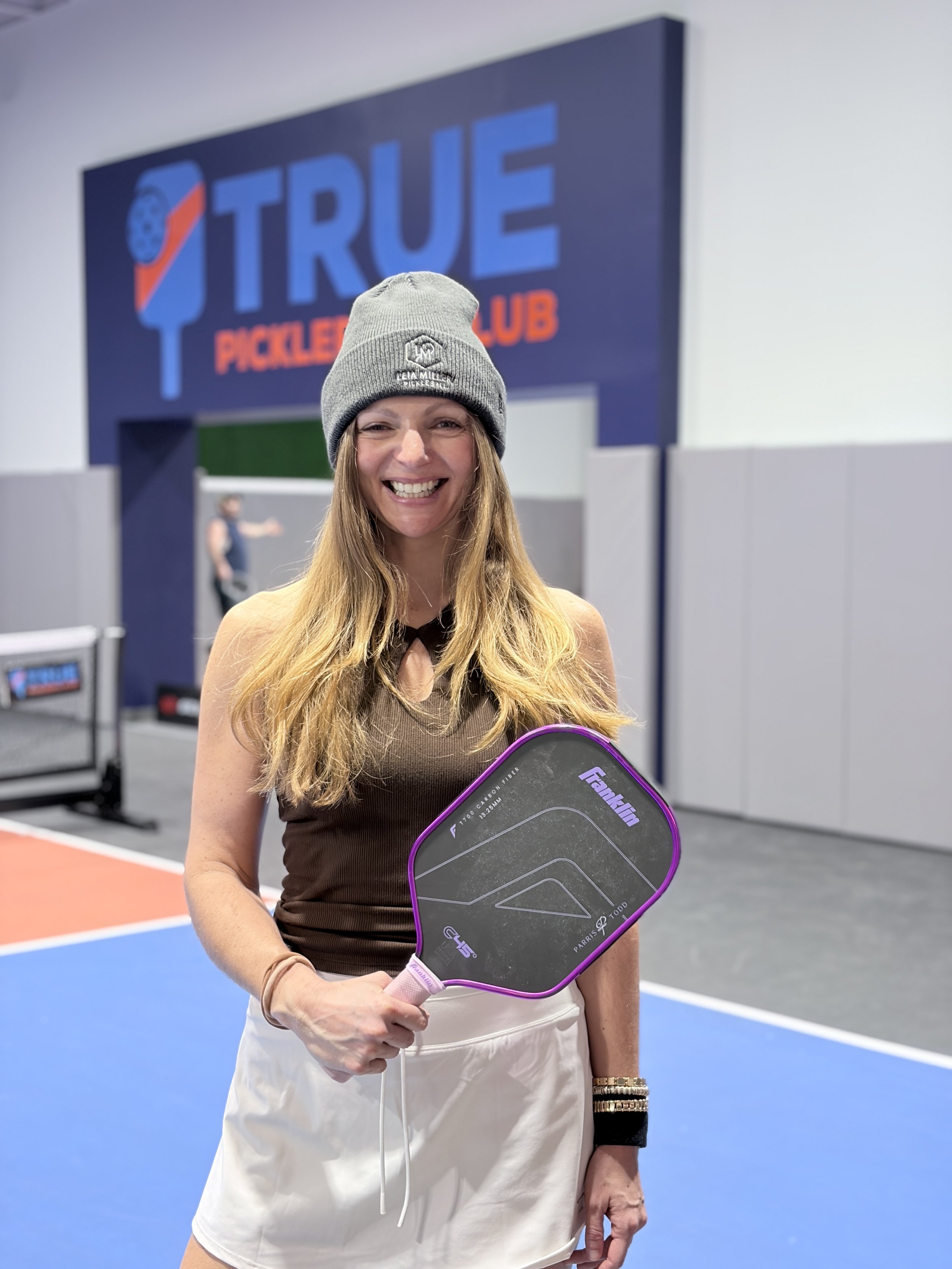 A smiling woman in athletic wear holding a pickleball paddle on a pickleball court.